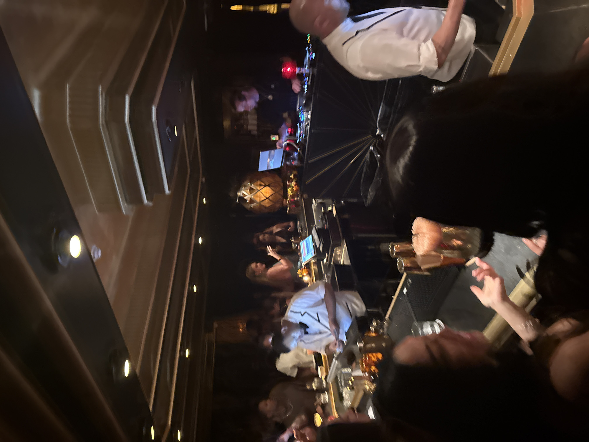 The image depicts a crowded bar scene, likely in the evening, with a focus on a DJ setup and patrons enjoying drinks. The atmosphere appears lively and dimly lit.
People:
Several people are present, including:
- A DJ: positioned behind a mixing console, wearing a white shirt.
- Bar patrons: seated or standing at the bar, some interacting with each other, with drinks in front of them.
Subjects:
- DJ equipment: a mixing console with various controls and screens.
- Drinks: various glasses containing beverages are visible on the bar.
Objects:
- Bar counter: a long, polished bar surface cluttered with drinks and other items.
- Bar stools: chairs for patrons to sit at the bar.
- Lighting: spotlights are embedded in the bar's architecture.
Actions:
- Patrons are ordering and consuming drinks, listening to music.
- The DJ is operating the mixing console, likely playing music.
Interactions:
- Patrons are talking and gesturing to each other.
- There is an interaction between bartenders and patrons.
Environment:
- Interior: The scene appears to be inside a bar or club, with dark, modern décor.
- Lighting: The lighting is dim and atmospheric.
- Location: Based on the style and atmosphere of the image, it is possibly located in Miami Beach, United States.