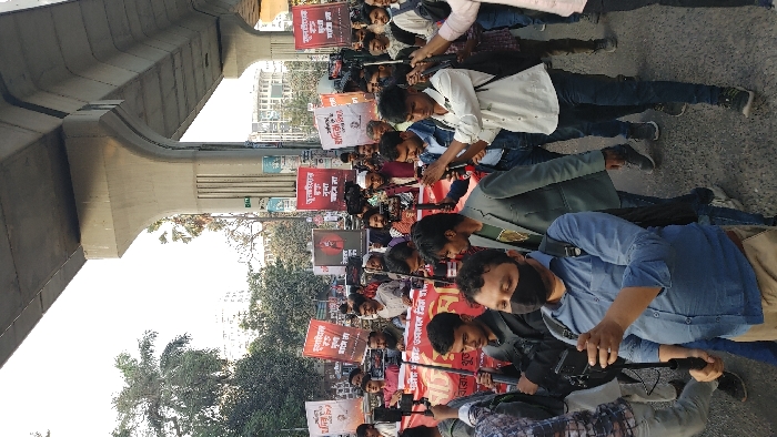 A daytime image captures a large gathering of individuals, primarily male, participating in a street procession or protest in Dhaka, Bangladesh. The group occupies an asphalt road beneath a prominent elevated concrete viaduct, consistent with the Dhaka Metro Rail infrastructure.
Numerous participants are holding up rectangular placards, predominantly red and white, displaying text in Bengali script and some featuring printed images of individuals. A large red banner, also with Bengali text, is held horizontally across the crowd in the mid-ground. Many individuals within the procession are actively documenting the event; several are visible holding smartphones mounted on stabilization sticks or gimbals, pointing them forward.
In the immediate foreground, a man wearing a blue collared shirt, khaki pants, and a black face mask is operating a smartphone attached to a handheld stabilizer. Other individuals are dressed in varied casual attire. In the mid-ground, one man is identifiable by a white skullcap and a maroon long-sleeved shirt.
The elevated concrete structure features robust support pillars. In the background, beyond the marching crowd, scattered palm trees and other greenery are visible, interspersed with multi-story urban buildings under a clear sky.