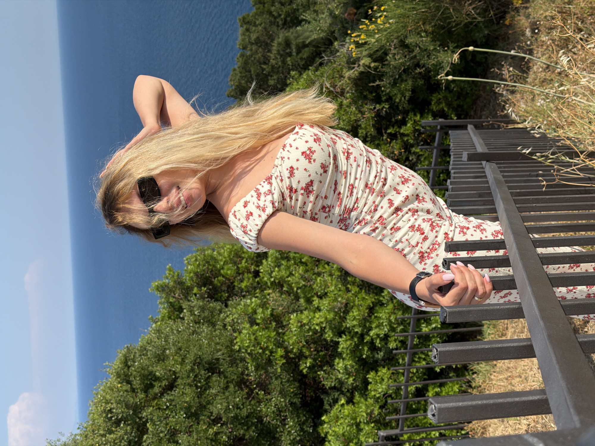 In Budva, Montenegro, a young woman with long blonde hair and sunglasses stands on a paved staircase overlooking the Adriatic Sea. She is wearing a white floral print dress and is leaning against a black metal fence. Her left hand is raised to her head, and her right hand is holding a small black object. Lush green trees and bushes surround the staircase, and the sea stretches out to the horizon under a clear blue sky.