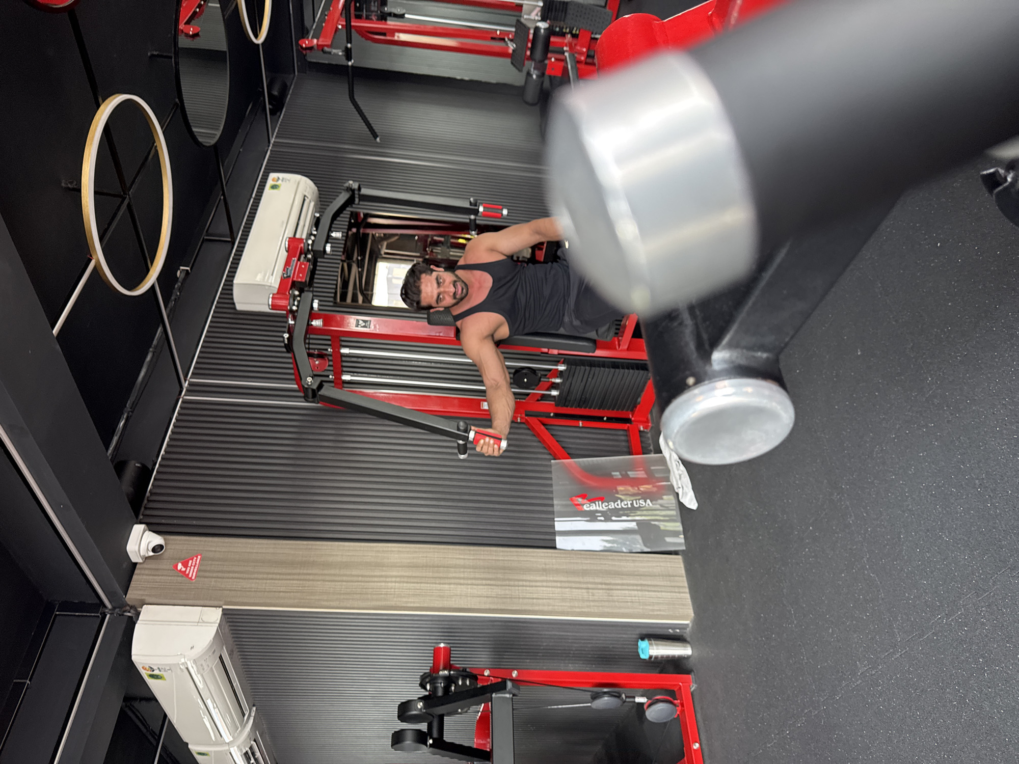 A man in a black tank top is using a chest fly machine in a gym. The gym has black walls with horizontal corrugated metal panels. There are mirrors, ring lights, and air conditioning units on the walls. Other gym equipment is visible in the background. A "ealleader USA" logo is visible on a piece of equipment. The location is not indicated in the image. Kolkata, India, is not indicated in the image.