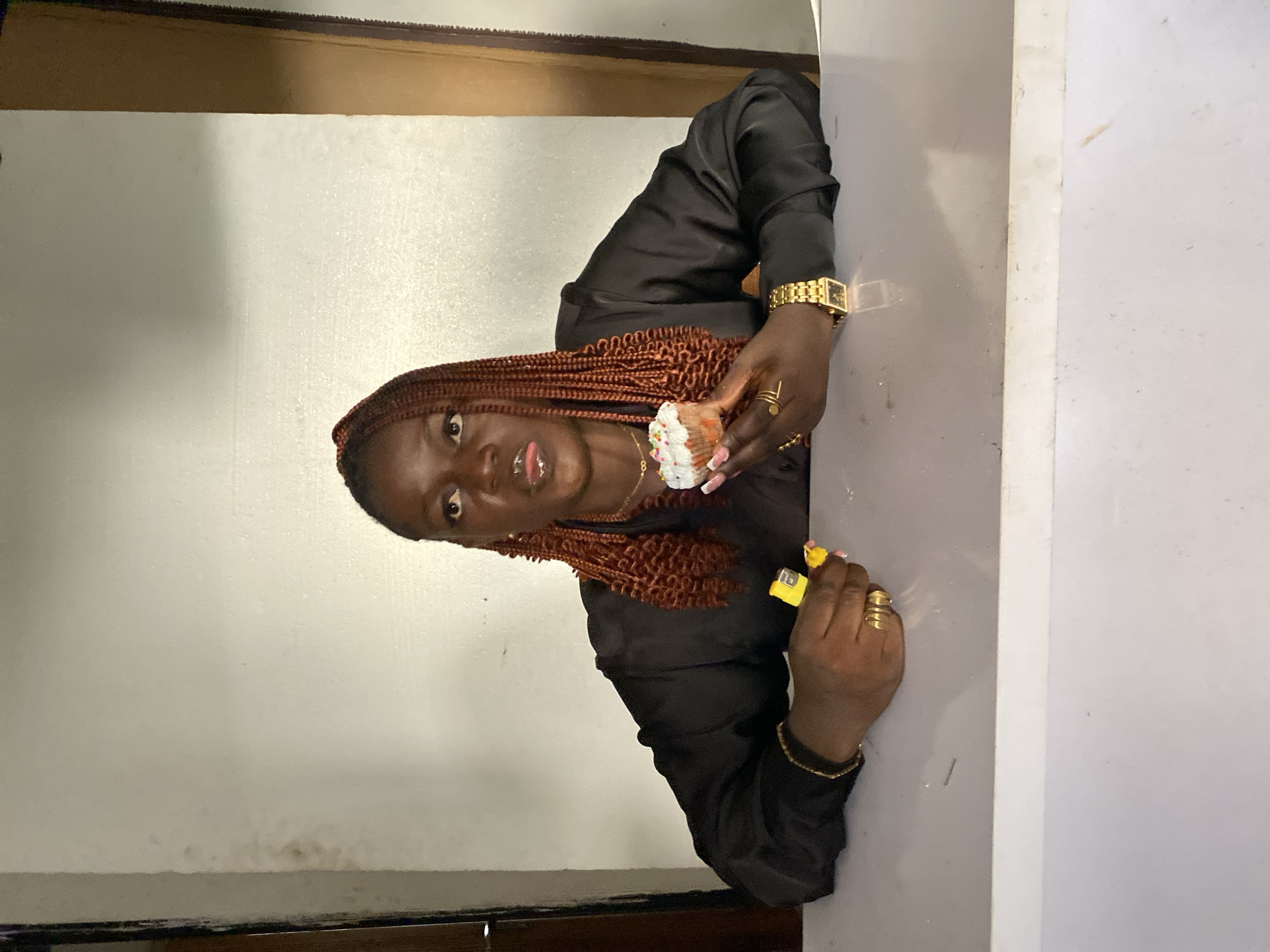 A brown-skinned woman with braided hair stands in what appears to be a building in Bukuru, Nigeria. She is holding a cupcake with white frosting and sprinkles in one hand and a yellow cigarette lighter in the other. She wears a black satin shirt, a gold watch, a necklace, and rings. The wall behind her is a light shade of gray and has some minor stains. There is a wooden trim at the top of the wall. The lighting in the scene is dim.