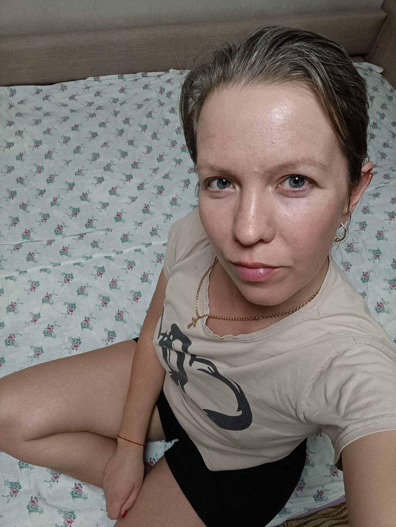 A woman sits cross-legged on a bed with a floral-patterned white bedsheet. She is wearing a light beige t-shirt with a black graphic design, and black shorts. A gold chain and a small gold cross are visible around her neck. She is also wearing gold earrings. She has fair skin, blue eyes, and grey hair. She is looking directly at the camera with a neutral expression. The headboard is beige. The scene is likely indoors and based on the user's location, potentially in the city of Chaikovsky, Russia.