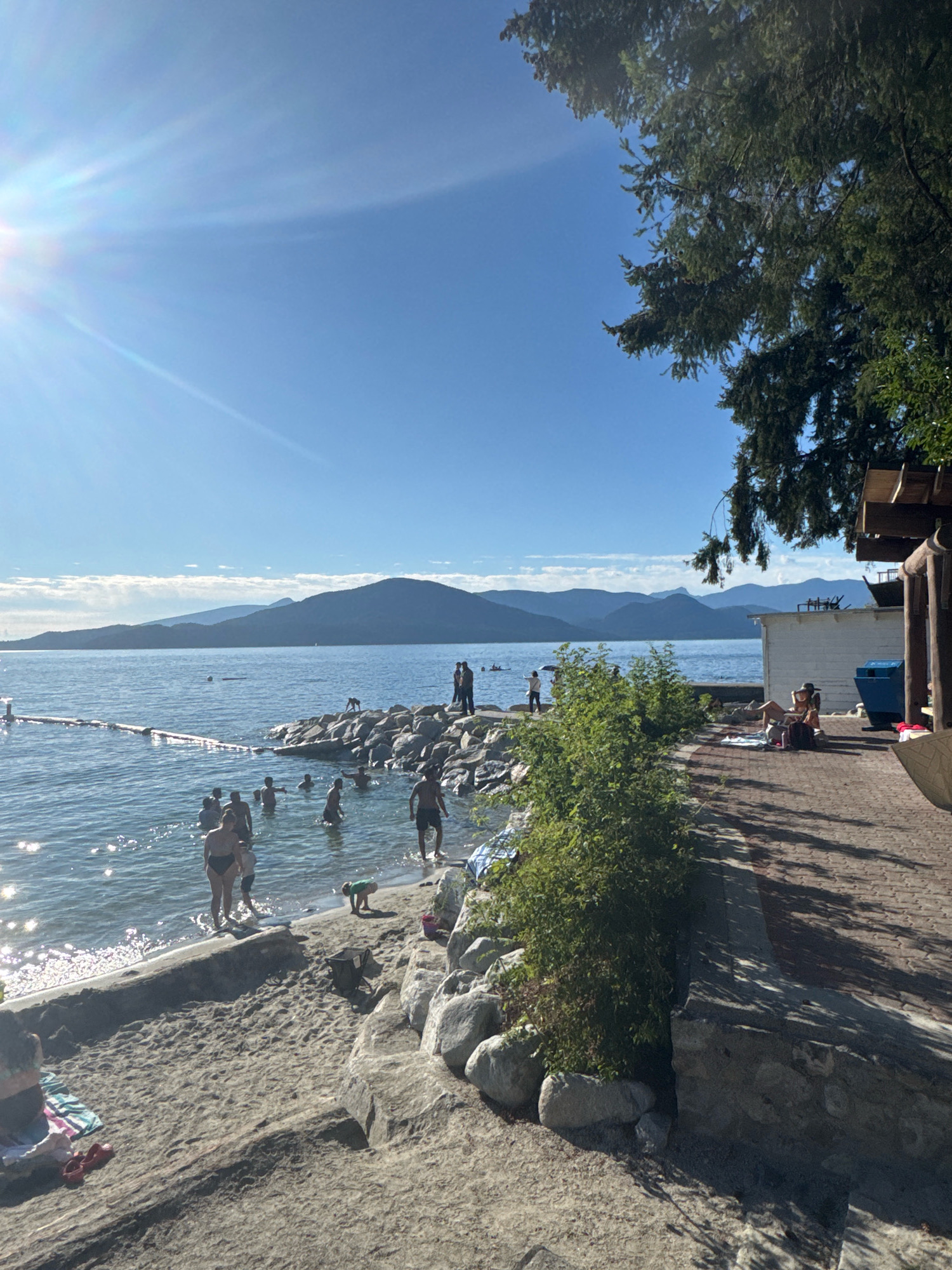 This image depicts a sunny day at a beach in Lions Bay, Canada. In the foreground, a sandy beach slopes down towards the water, with some rocks and vegetation along the edge. Several people are visible on the beach, some lying on towels and others standing.
The water is calm and clear, with a rope barrier marking a swimming area. A group of people are wading and swimming in the water. In the background, a rocky shoreline extends into the water, with more people gathered on the rocks. Beyond the water, a range of mountains forms the horizon under a clear blue sky.
To the right of the image, a paved walkway runs along the shore, lined with trees and a building. A few people are seated on the walkway, enjoying the view. The overall scene is one of a recreational area with people engaging in various activities on a warm, sunny day.