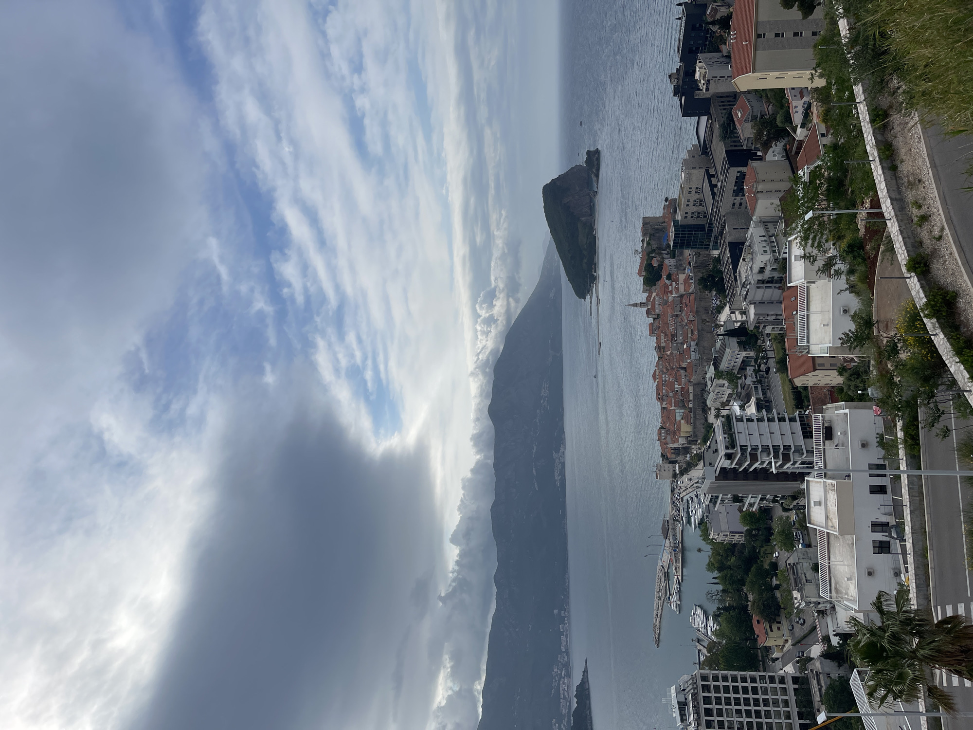 The image captures a vertical aerial view of Budva, Montenegro, encompassing the Adriatic Sea, a portion of the city, and the surrounding mountainous terrain under a partly cloudy sky.
The foreground shows the coastline with a road lined with trees and street lights, adjacent to modern buildings with white facades. Closer to the sea, there's a harbor area filled with numerous yachts and boats. The older part of Budva, with its characteristic red-tiled roofs, extends along the coast.
In the mid-ground, the Adriatic Sea dominates, leading the eye towards a small rocky island and the base of a large, shadowed mountain range. A bank of dark clouds covers a portion of the sky behind the mountains.
The sky is predominantly blue, mixed with white cirrus clouds and denser gray cloud formations. The image is taken from an elevated position, likely from a high-rise building or a mountain vantage point.
