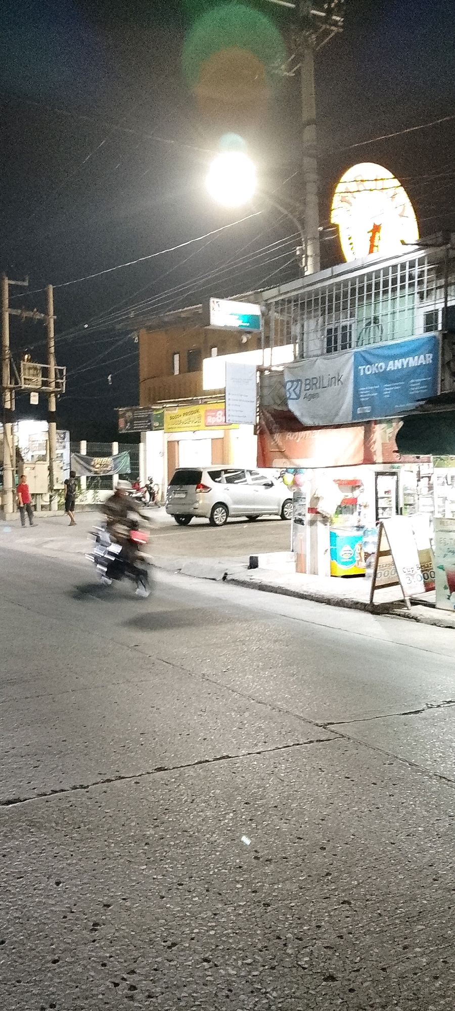 This is a night-time, outdoor, low-light photograph taken on a street in Satriamekar, Indonesia. The scene depicts a street with a moving motorcycle in the foreground, creating motion blur. The motorcycle rider is wearing dark clothing and a hat. To the right of the motorcycle, a silver minivan is parked. Further to the right, there is a storefront with a blue sign that reads "BRIlink" and "TOKO ANYMAR". There are other signs and banners visible, including one with red lettering and a white background that appears to be a price advertisement. In the background, there are buildings, utility poles with numerous wires, and streetlights. Two individuals are standing on the sidewalk to the left of the motorcycle. One person is wearing a red shirt, and the other is wearing dark clothing. The road surface is asphalt with visible cracks and variations in texture. The overall lighting is dim, with bright streetlights casting light and creating lens flares.
