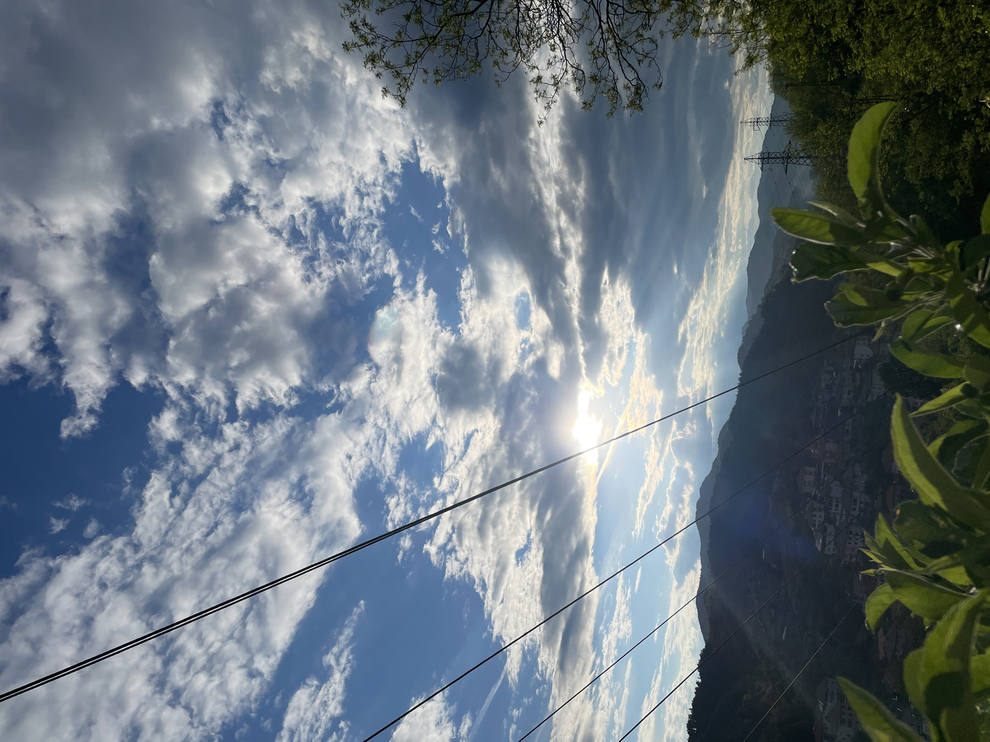 The image presents a landscape view, likely taken in Zlatograd, Bulgaria, given the mountainous terrain and architectural style. The sky dominates the frame, filled with a mix of cumulus and stratus clouds, interspersed with patches of blue. The sun is visible, partially obscured by clouds, creating a bright glare. In the lower portion of the image, a hillside is seen, dotted with buildings that suggest a densely populated area. Green foliage, possibly trees or bushes, frames the scene on the right and top, partially obscuring the view. Power lines stretch across the foreground, adding a linear element to the composition. In the middle ground, two lattice towers are visible on the hill, indicating the presence of electrical infrastructure. The lighting suggests it is either early morning or late afternoon, given the low angle of the sun.