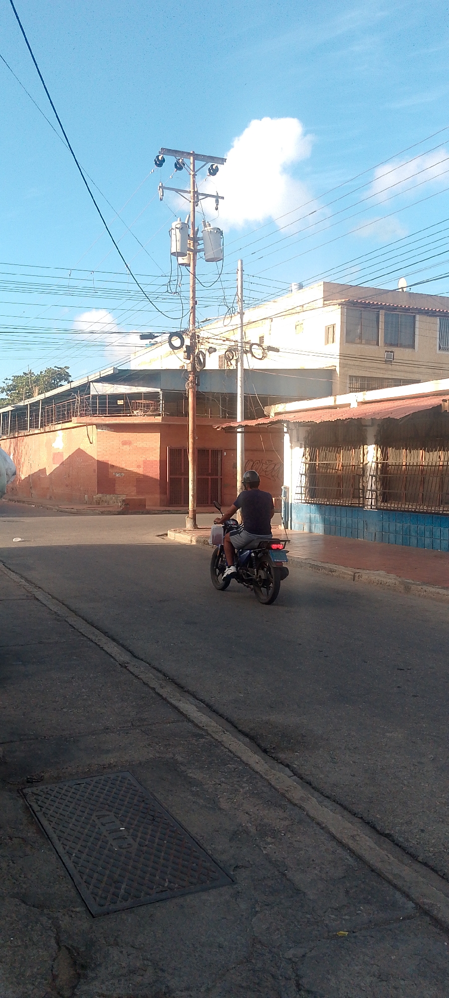 A man wearing a dark t-shirt and shorts rides a motorcycle down a street in Juan Griego, Venezuela. The motorcycle is dark in color with a red taillight and a license plate. He is wearing a dark helmet. The street appears to be paved with asphalt, with a white line marking the center. A manhole cover with a diamond pattern is visible on the left side of the road. On the right side of the street, there are buildings with orange brick walls and metal bars on the windows. A power pole with electrical transformers and wires is also present. The sky is blue with some white clouds.
