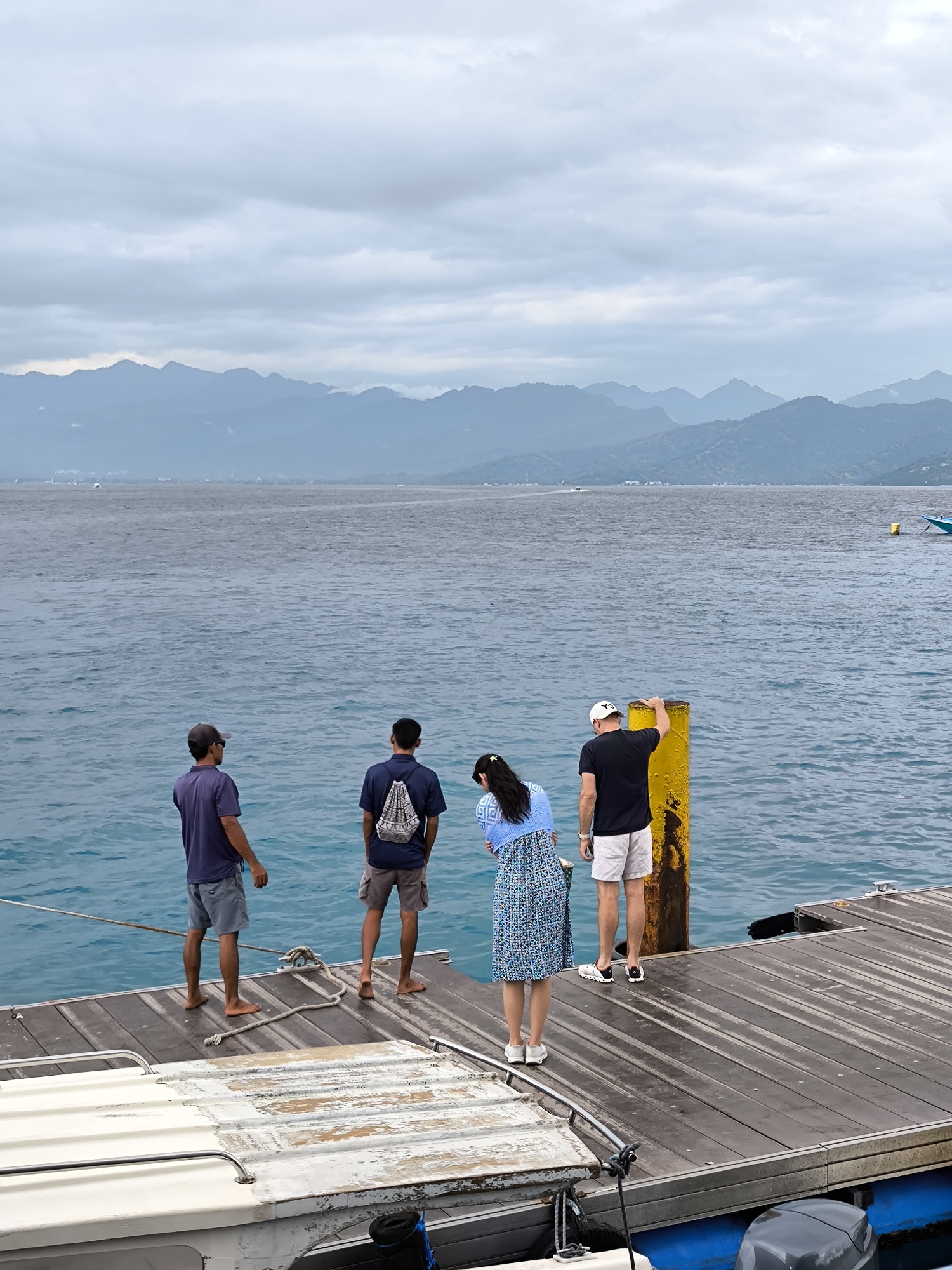 Four people stand on a wooden pier overlooking a body of water, likely the sea. From left to right, the first man wears a dark-colored baseball cap, a blue short-sleeved shirt, and gray shorts. He is barefoot. Next to him, the second man has a dark blue short-sleeved shirt, gray shorts, and a white patterned backpack. He is also barefoot. The third figure is a woman with long dark hair. She wears a light blue patterned dress, a matching cardigan, and white sneakers. The fourth person, a man, is dressed in a black short-sleeved shirt, light-colored shorts, a white baseball cap, and dark-colored sneakers. He rests his hand on a yellow cylindrical post.
The pier is made of dark, wet wooden planks. In the foreground, a white boat with a gray motor is visible. A thick rope lies coiled on the pier.
In the background, a range of mountains is seen under a cloudy sky. The water is a deep blue color. The location is identified as Unknown, Indonesia.