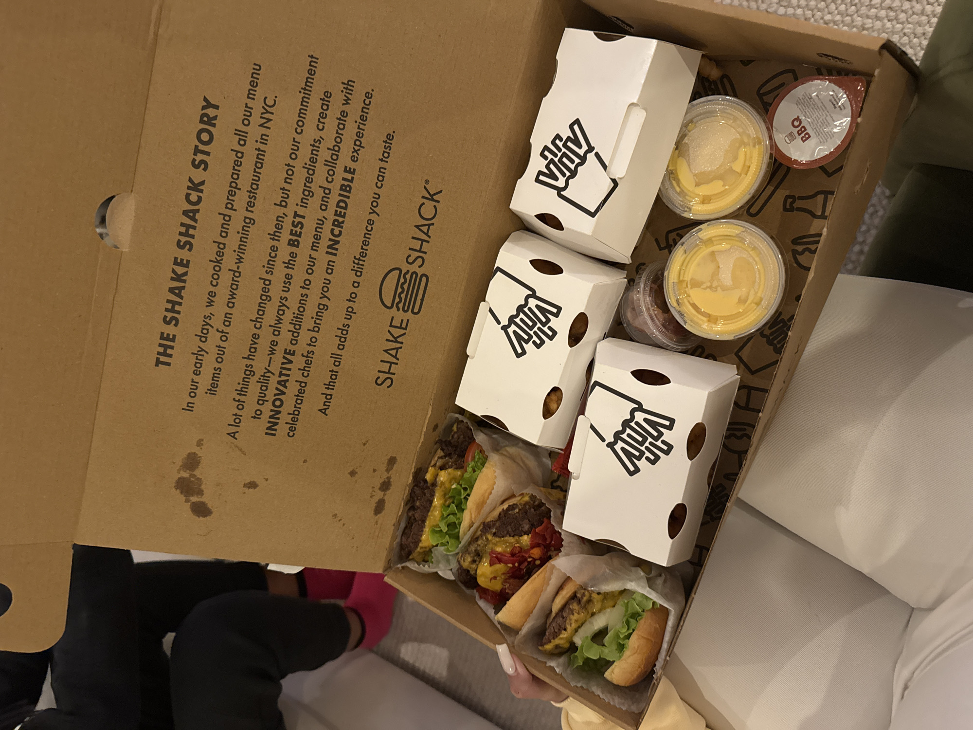 An overhead shot shows a Shake Shack meal delivered in a branded brown cardboard box, likely indoors in London, United Kingdom. The box contains three unwrapped burgers with patties, cheese, lettuce, and buns, along with three small white cartons featuring a black icon resembling a hand holding fries, suggesting they contain fries. Two clear plastic cups filled with a yellow liquid, possibly cheese sauce, and a small red container labeled "BBQ" are also present.
The inside lid of the box displays "THE SHAKE SHACK STORY," a text detailing the brand's commitment to "BEST ingredients," "INNOVATIVE" menu additions, and an "INCREDIBLE experience." The Shake Shack logo, including a burger graphic, is prominent.
A person's legs, dressed in dark clothing with bright pink socks, are visible in the bottom left, implying the box is on a lap or low surface. Another person's hand is visible on the right, holding one of the burgers. A light-colored, soft furnishing, possibly a couch or chair, fills the right side of the frame, contributing to a casual, indoor setting.
