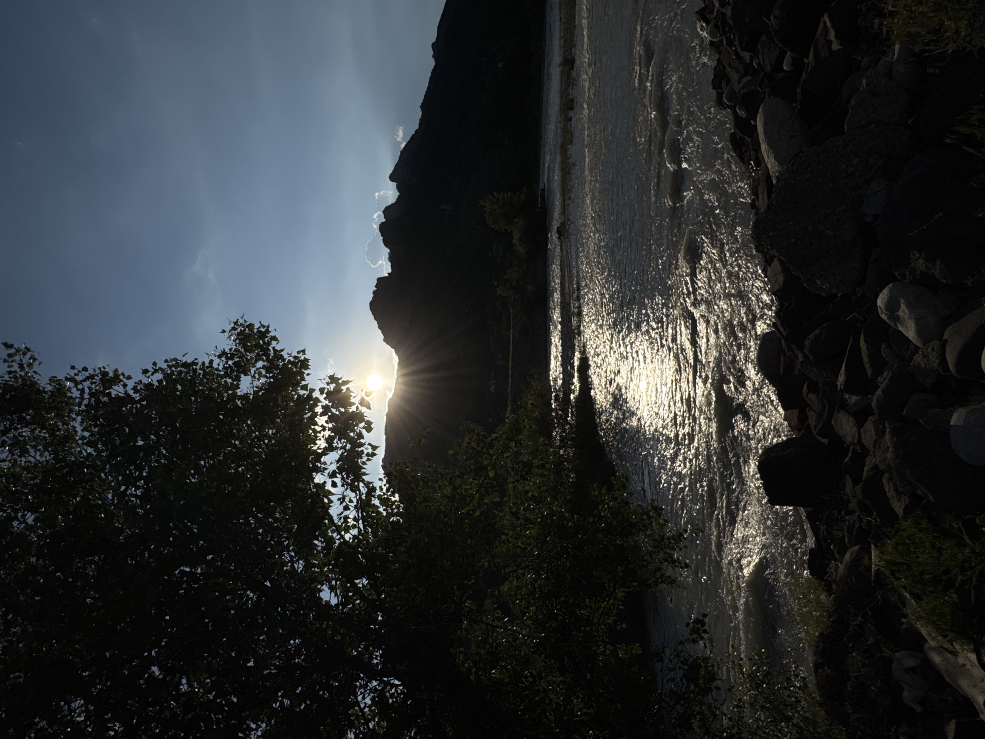The image depicts a natural scene at sunset, likely near the city of Karachayevsk, Russia, given its mountainous terrain and river. A large, silhouetted tree dominates the left foreground, its dark foliage obscuring part of the sky and the setting sun. The sun, partially hidden behind a jagged mountain range in the background, casts a bright glare and visible sunburst rays across the sky. The water in the foreground is a river, its surface shimmering with reflected sunlight, indicating movement and a slightly choppy texture. The riverbanks are lined with dark, rough-textured rocks of varying sizes. In the distance, the silhouetted mountain range forms a dramatic backdrop against the clear blue sky, with a few wispy clouds visible near the mountain peaks. The overall atmosphere is serene and natural, with the low angle of the sun suggesting either early morning or late evening. There are no people or man-made structures clearly visible in the image, emphasizing the wild and untouched quality of the environment.