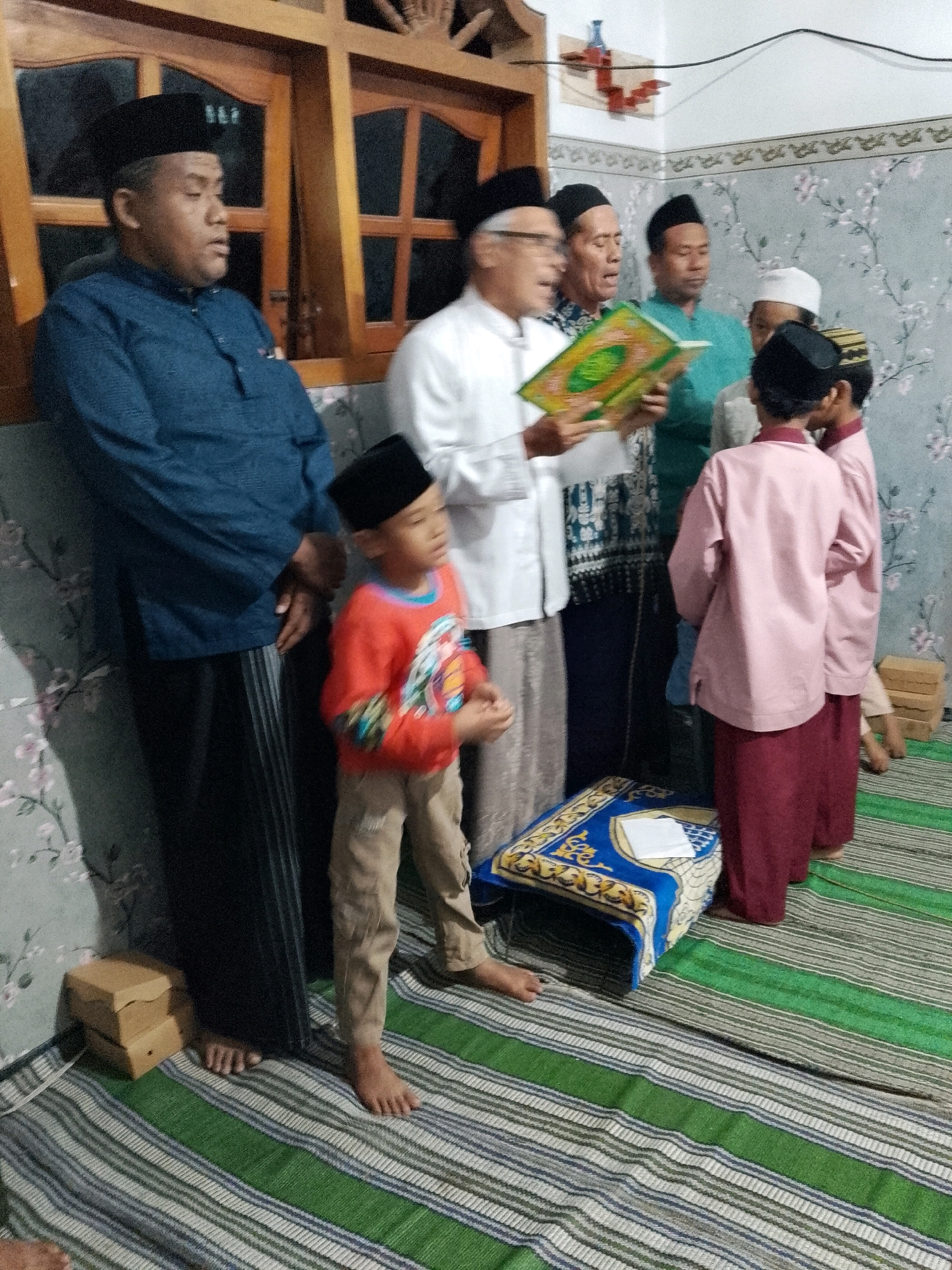 A group of at least seven males, ranging from children to adults, are gathered indoors. The men appear to be engaged in a religious activity, with one adult male holding an open book, possibly a religious text, and others looking in his direction. The environment is an indoor setting, with a floral patterned wallpaper on the wall and a wooden window frame visible. The floor is covered with a striped mat. Several objects are present, including what appear to be small boxes stacked in a corner, and a prayer mat on the floor. The people are dressed in traditional attire, including caps and long garments. The lighting suggests an indoor scene, possibly during the evening or a dimly lit room. The overall impression is that of a religious gathering or ceremony. This scene is located in Rejoyoso, Indonesia.