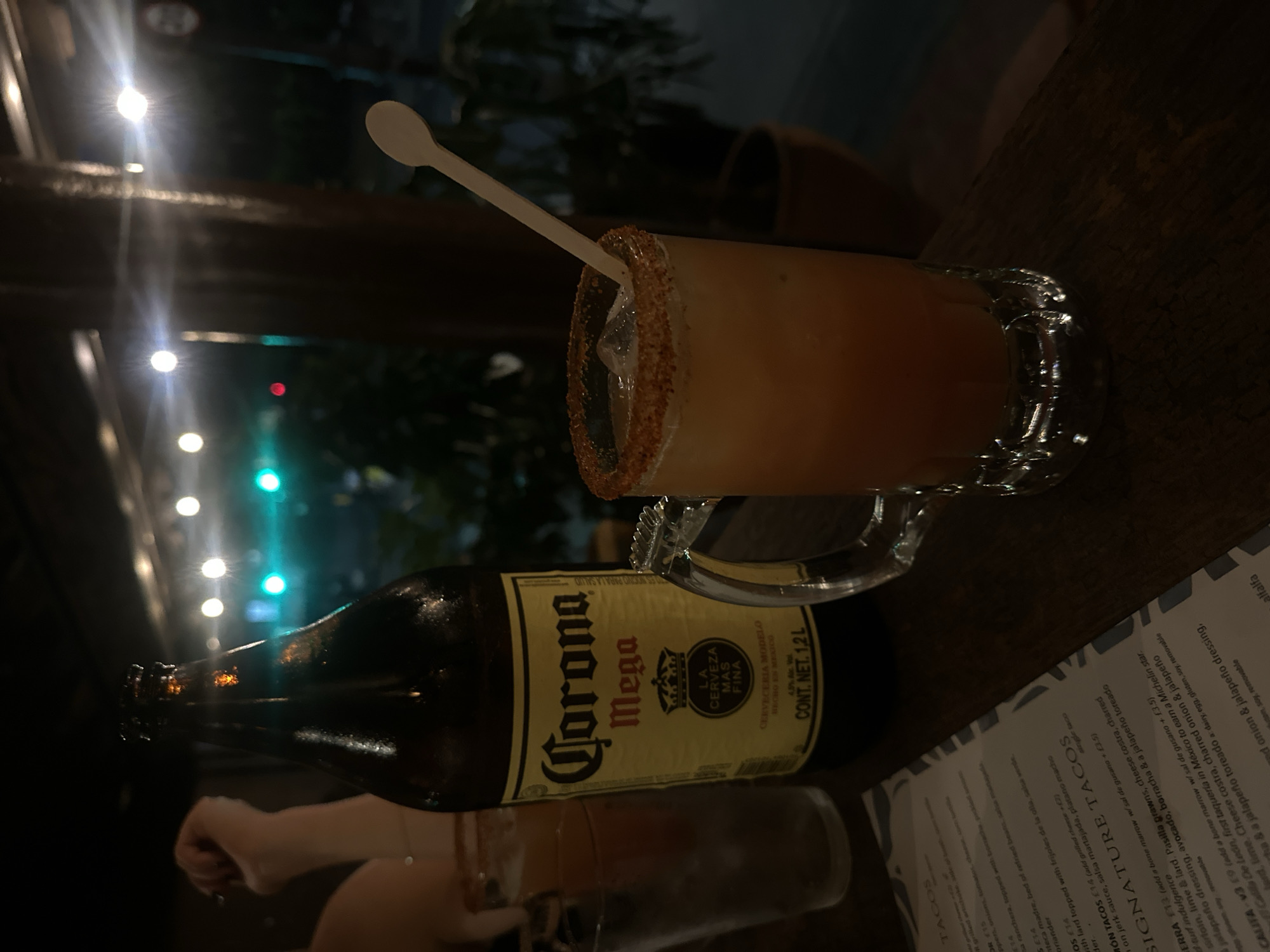 The image shows a table setting with a Corona Mega beer bottle, a glass of beer, and a menu. The glass has a salted rim and a small plastic spoon. The menu is titled "SIGNATURE TACOS". Based on the text on the menu and the general presentation, it appears to be a restaurant setting. The image does not contain sufficient information to establish the location as London, United Kingdom.