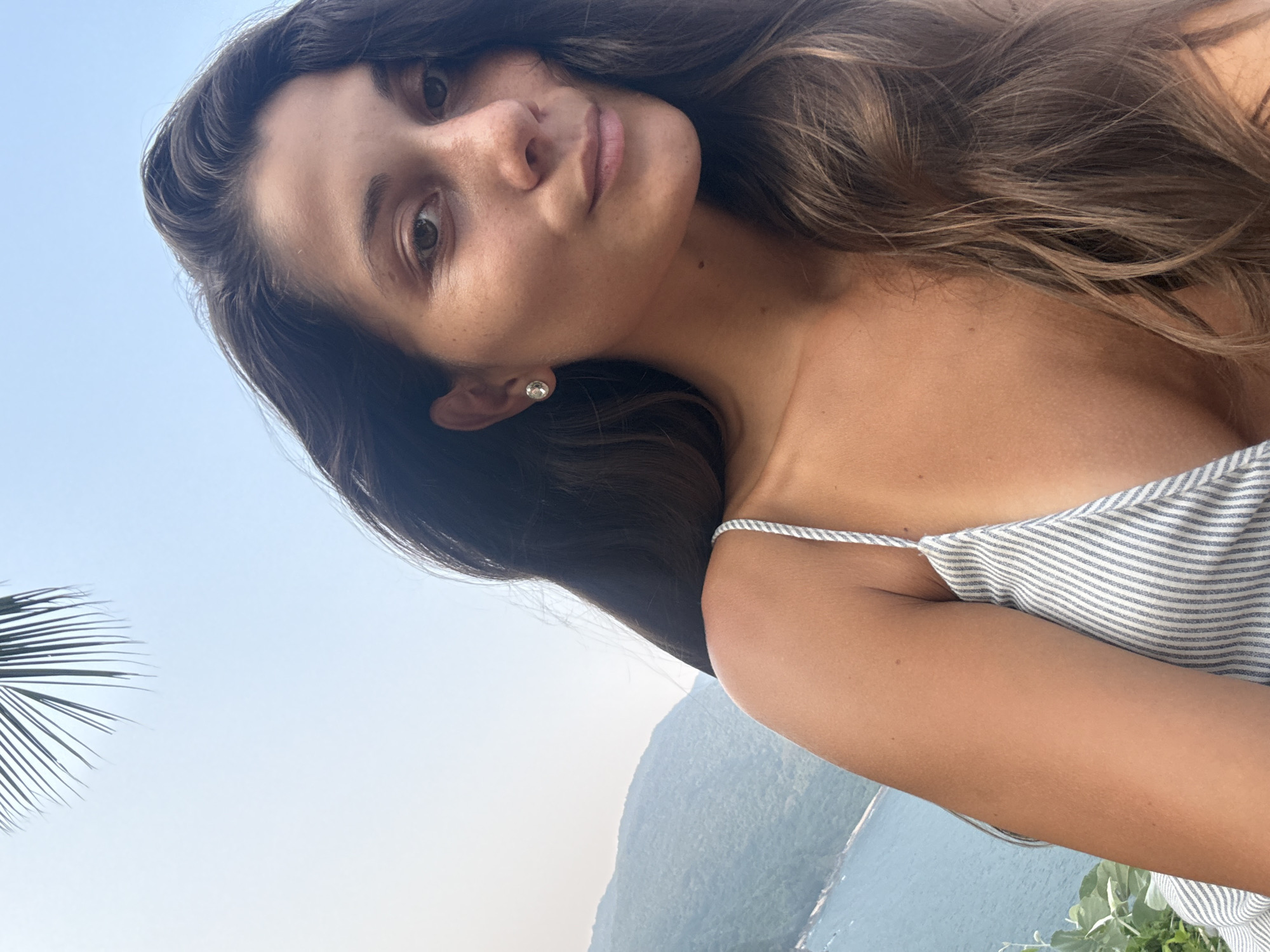 A woman poses for a selfie outdoors. Her dark hair is long and wavy, framing her face, and she wears a light-colored, striped top with thin straps. A small, diamond-like earring is visible on her left ear. The background features a body of water, possibly the ocean, and mountains, suggesting a coastal location. A palm frond is partially visible in the lower left corner. Given the location query, this may be a view of the ocean near Đà Nẵng, Vietnam.