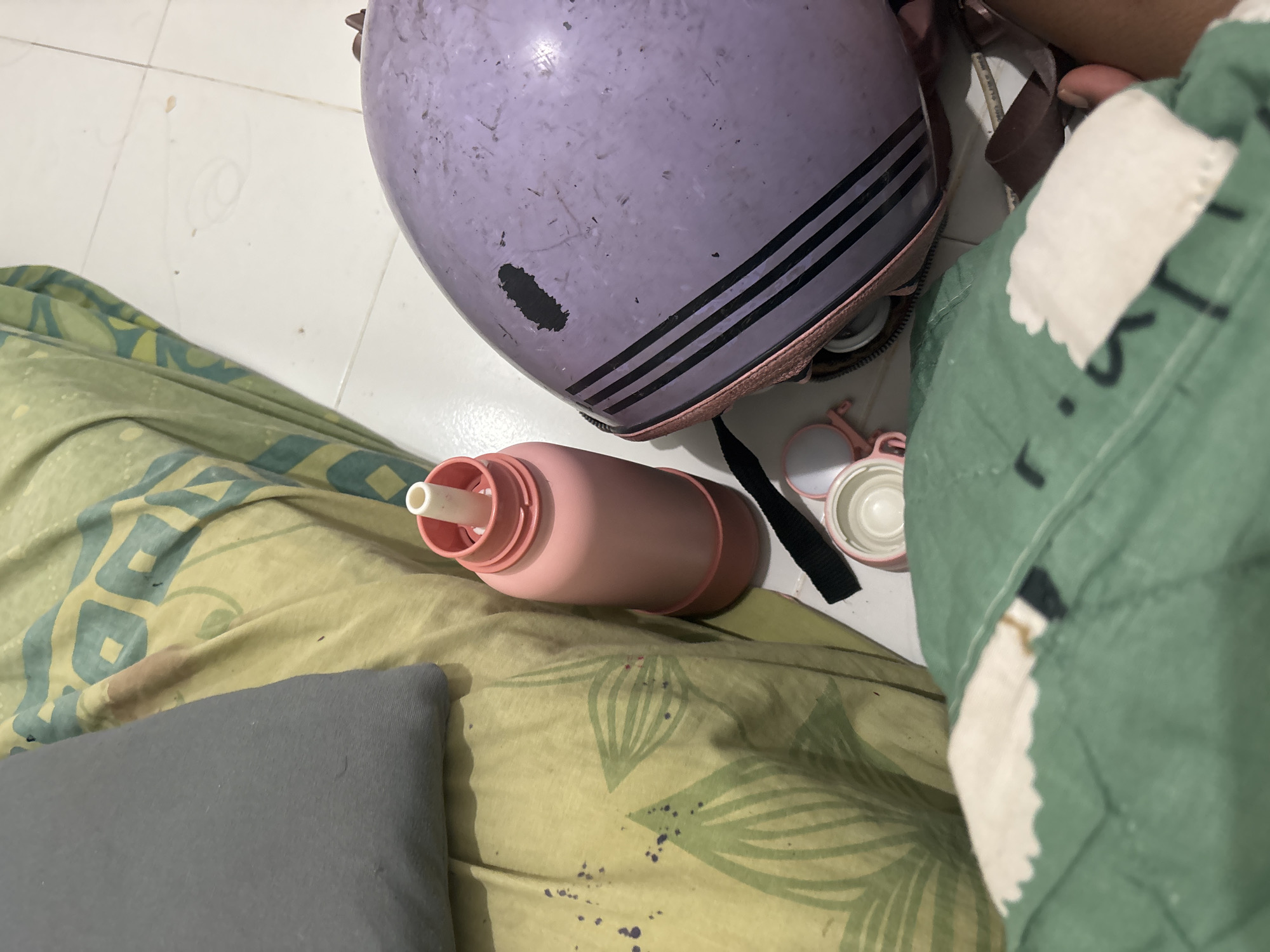 The image depicts a domestic scene within a room, possibly a bedroom, in North Jakarta, Indonesia. The floor is tiled with white squares. On the floor, a light purple helmet with black stripes is visible, along with a pink water bottle and a small pink accessory. A portion of a green blanket with a white pattern is in the foreground, and a yellow and green patterned sheet covers a surface, likely a bed, on the left side of the image. A gray pillow is also present on the left. The overall impression is of a casual, everyday setting.