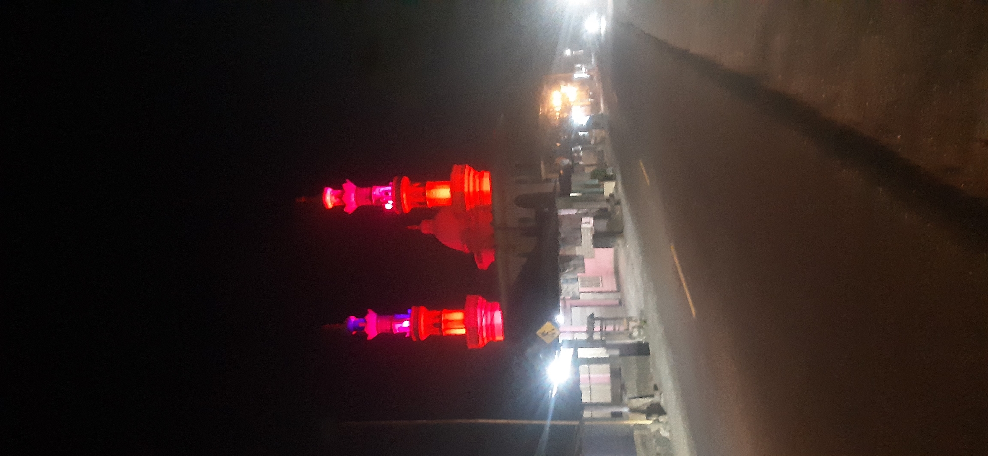 The image depicts a night scene in Sepakek, Indonesia, focusing on a mosque illuminated with red and purple lights. Two prominent minarets, glowing intensely with red light and accented with purple at their peaks, flank a central dome structure of the mosque. The mosque appears to be situated along a paved road, with a street light visible on the left side of the road casting a bright white glow. Adjacent to the mosque, on the right side of the road, is a row of low-rise buildings or shops with illuminated windows and awnings. The road itself is asphalt, with a yellow line marking the center. The overall atmosphere is dark, with the artificial lights from the mosque and buildings creating the primary illumination. The image is slightly blurred, suggesting movement or low light conditions during capture. There are no people or vehicles clearly visible in the immediate foreground or around the mosque.