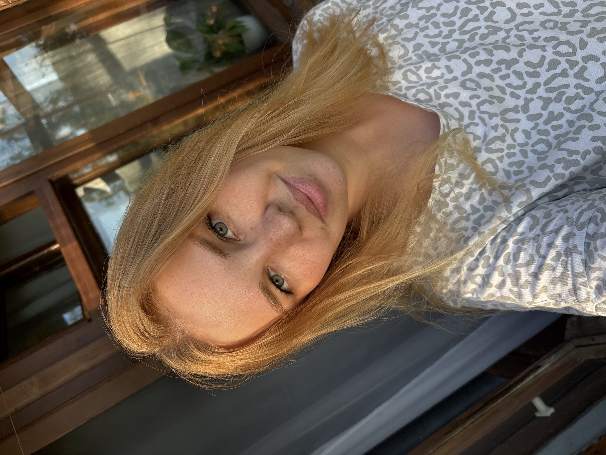 A young woman with blonde hair and green eyes, freckled cheeks, and light pink lipstick smiles at the camera in a close-up selfie. She is wearing a grey and white leopard-print t-shirt. The background suggests an outdoor setting with a wooden window frame and some greenery visible through the glass. The lighting indicates it is likely late afternoon or early morning, with warm sunlight illuminating the woman's face and hair. The overall impression is casual and natural.