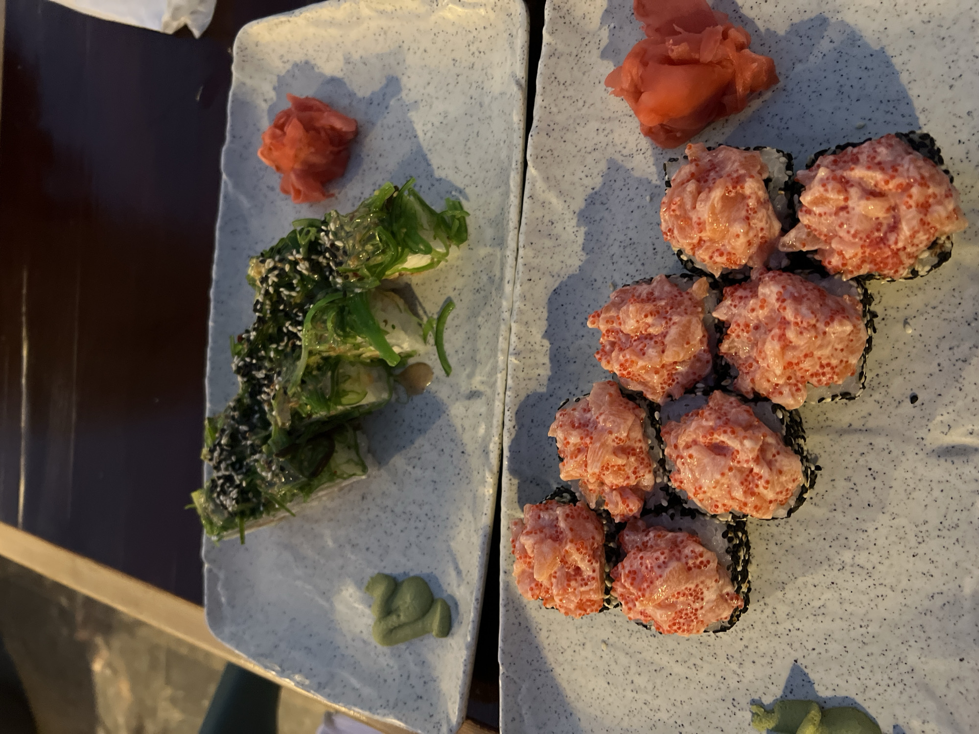 Two plates of sushi are shown. The top plate contains several rolls topped with seaweed and sesame seeds, served with a side of pickled ginger and wasabi. The bottom plate displays several rolls topped with a creamy pink sauce and red roe, also served with pickled ginger and wasabi. The plates are placed on a dark, wooden table. The background is out of focus, showing what appears to be an outdoor seating area with ambient lighting. No people are visible in the image. This scene is characteristic of a sushi restaurant in Constanța, Romania, given the typical offerings and presentation of such establishments in the city.