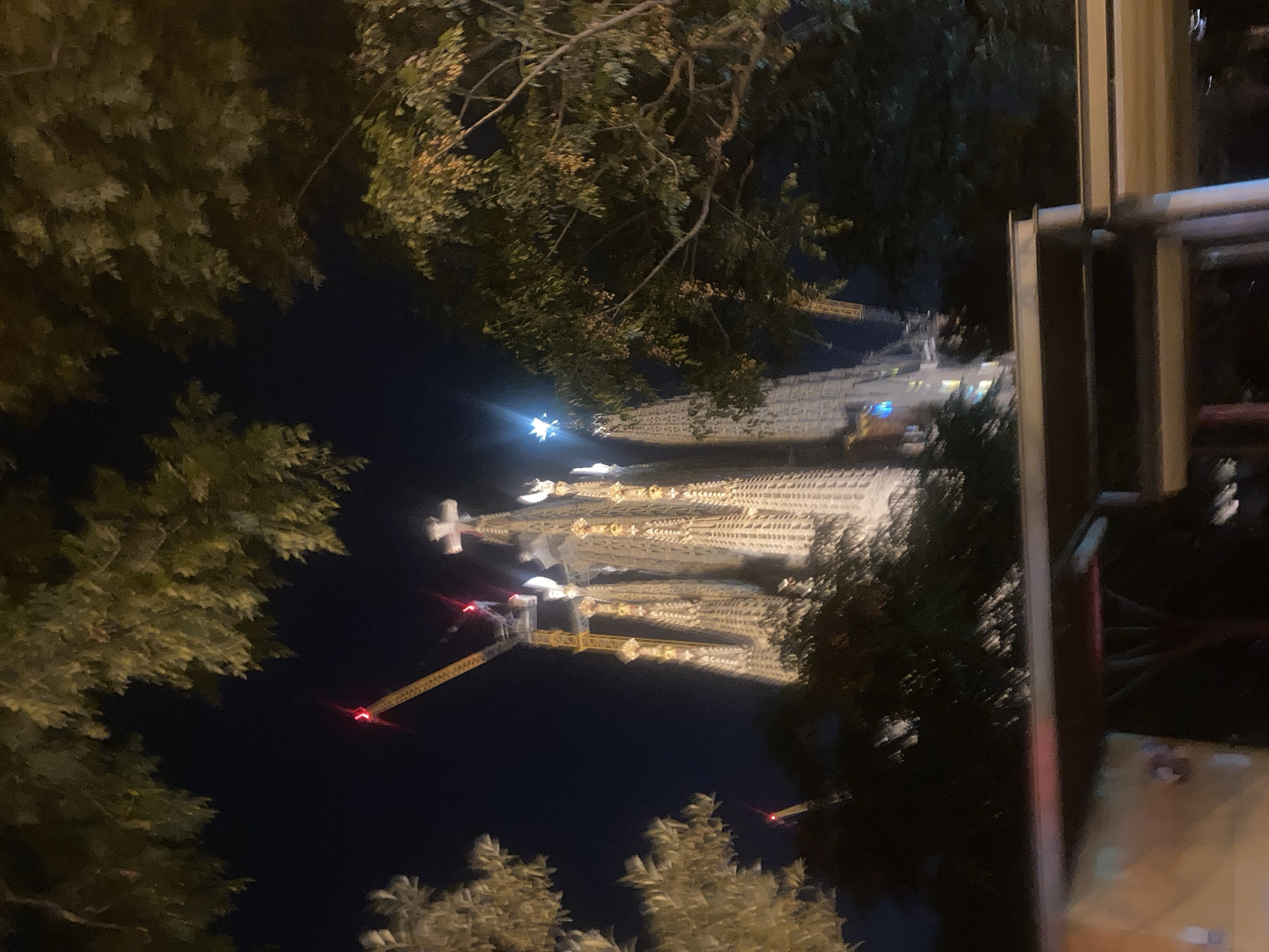 The image depicts the Sagrada Familia in Barcelona at night, viewed through trees and a partial railing. The Basilica is brightly lit and stands tall. Construction cranes are visible near the building. Trees obscure parts of the scene. The sky is dark.