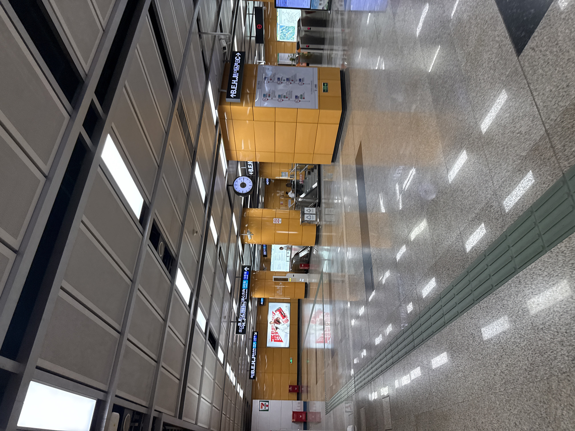 The image depicts the interior of a modern subway station, likely located in Haizhu District, China. The station features a clean and contemporary design with a polished, reflective floor that shows numerous bright light reflections. The ceiling is a modular system with integrated lighting panels.
Prominently visible are large, bright orange structural columns that serve as supports and display information. One such column in the foreground has a diagram on it, possibly a station map or route information. Above the columns, directional signs with both Chinese characters and English text are mounted on the ceiling. These signs indicate exits and directions to different lines, such as "B.E.H." and "Line 18".
In the background, escalators are visible, suggesting access to different levels of the station. There are also ticket gates and information kiosks. A large advertisement screen is mounted on an orange wall, displaying a vibrant image. A Seven Eleven convenience store is also identifiable by its signage on the left side of the image.
Several people are present, though mostly in the background and not the primary focus. One person is seen near the escalators, and others are in the distance, interacting with station amenities or waiting. The overall environment appears to be well-lit, clean, and organized, typical of a functional urban transportation hub. The flooring includes tactile paving for the visually impaired.