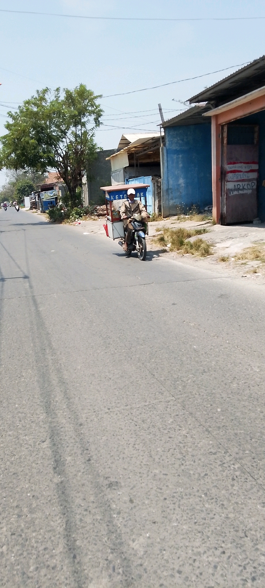 A man wearing a light-colored jacket and a white cap rides a motorcycle with a cart attached to the back. The cart is covered with a light brown awning and has a red bag hanging off the side. The man is on a paved road, which appears to be in a semi-urban or rural area. On the right side of the road, there is a blue building with a corrugated metal roof. Graffiti is visible on the building's entrance, with the words "WARUNG KOPI" and "WARKOP" written in red. A tree with green leaves is to the left of the road, and in the distance, more buildings and motorcycles are visible. The sky is clear and bright.