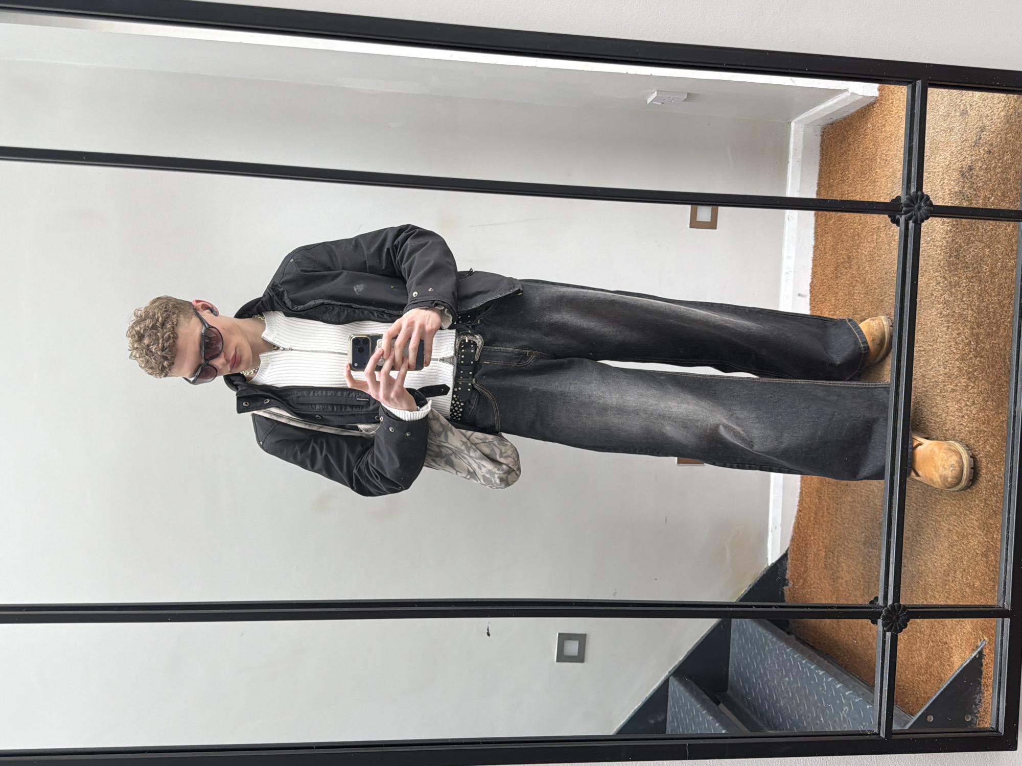 A young man is captured in a mirror selfie within an indoor environment. He sports a white, zip-up sweater layered under a dark jacket, complemented by wide-leg, black-wash jeans secured with a studded belt and tan boots. Accessories include dark sunglasses, a chain necklace and a patterned bag. He is holding a smartphone, presumably taking the photo. The background is a neutral-toned wall and a staircase. Based on the interior architecture and decor, the location is likely in a contemporary living space, possibly in the city of London, United Kingdom.