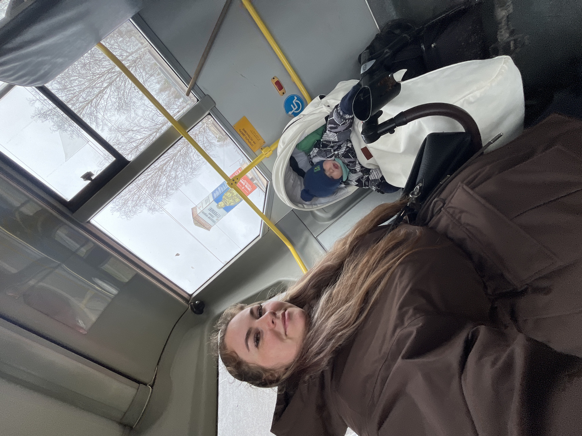 The image captures a selfie-style view of a woman and a young child inside a public transport vehicle, likely a bus, during the daytime in Chaikovsky, Russia.
In the foreground, a woman with long, light brown hair and a dark brown jacket looks towards the camera with a neutral to slightly pleasant expression. Behind her and to the right, a baby or young child is resting or sleeping in a white and black baby carriage. The child is wearing a blue winter hat and patterned winter clothing, appearing comfortable and calm.
The setting is the interior of a bus, characterized by light gray walls, yellow handrails, and large windows. Visible details inside the bus include a blue circular sign with a white wheelchair symbol, indicating accessibility features, and a yellow rectangular sign with partially visible Cyrillic text that reads "ОТКРОЙТЕ ДВЕРЬ" (Open the door).
Through the bus windows, the exterior view shows bare tree branches against an overcast, bright sky, suggesting a winter or early spring day. A sign or advertisement outside the bus, partially visible, also features Cyrillic text, possibly "ГЕРОЯМ!" (To heroes!). The overall impression is one of everyday travel on public transport, with the woman accompanying the child in their carriage.