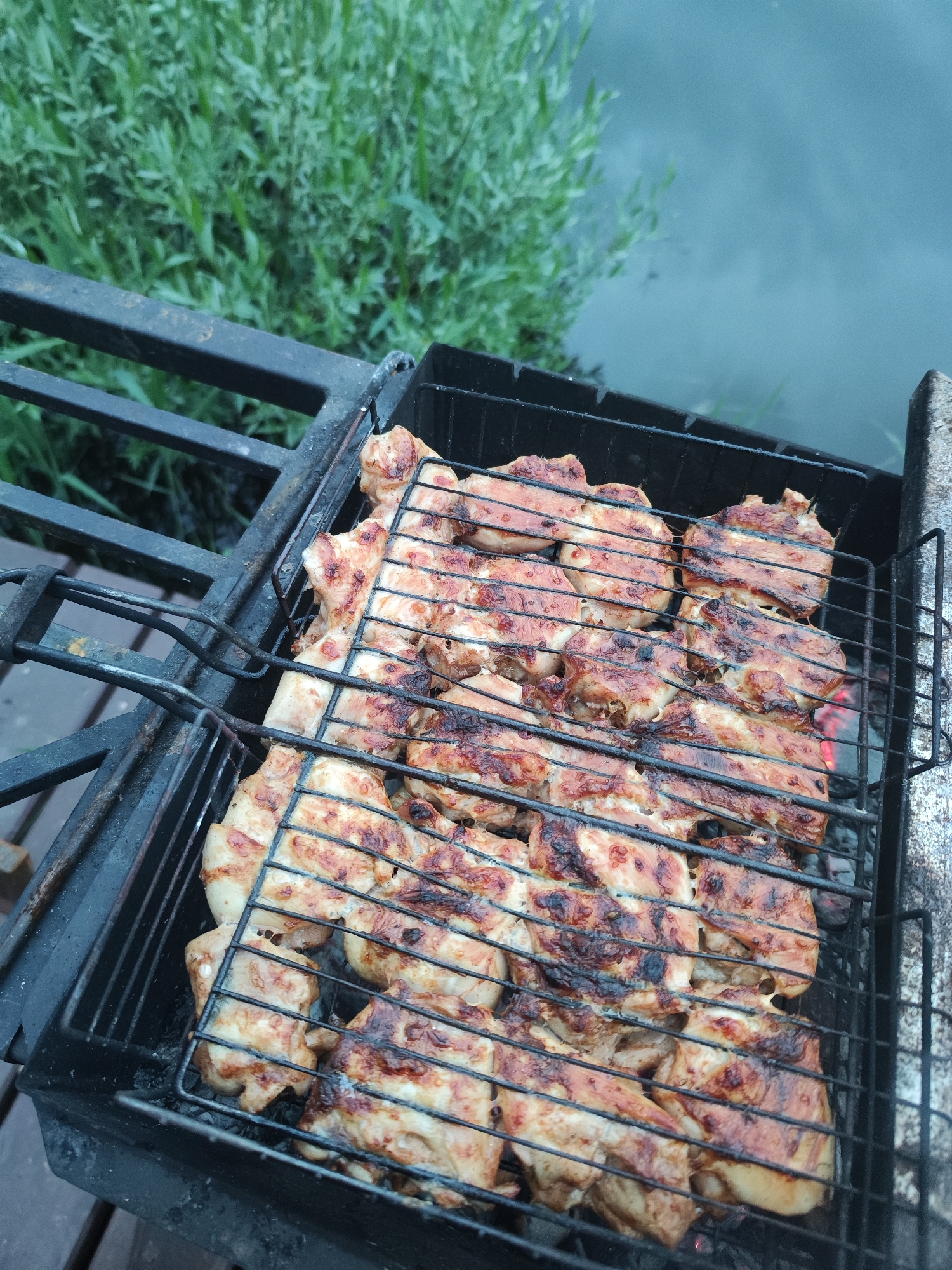 The image shows chicken being grilled on a charcoal grill. The grill is black and is located outdoors on a wooden deck, with a black metal railing to the left. In the background, there is green vegetation and what appears to be water, possibly a river or lake. The chicken pieces are arranged closely together on the grill and have grill marks, indicating they are being cooked. Based on visual evidence, the location is likely in or near Chaikovsky, Russia.