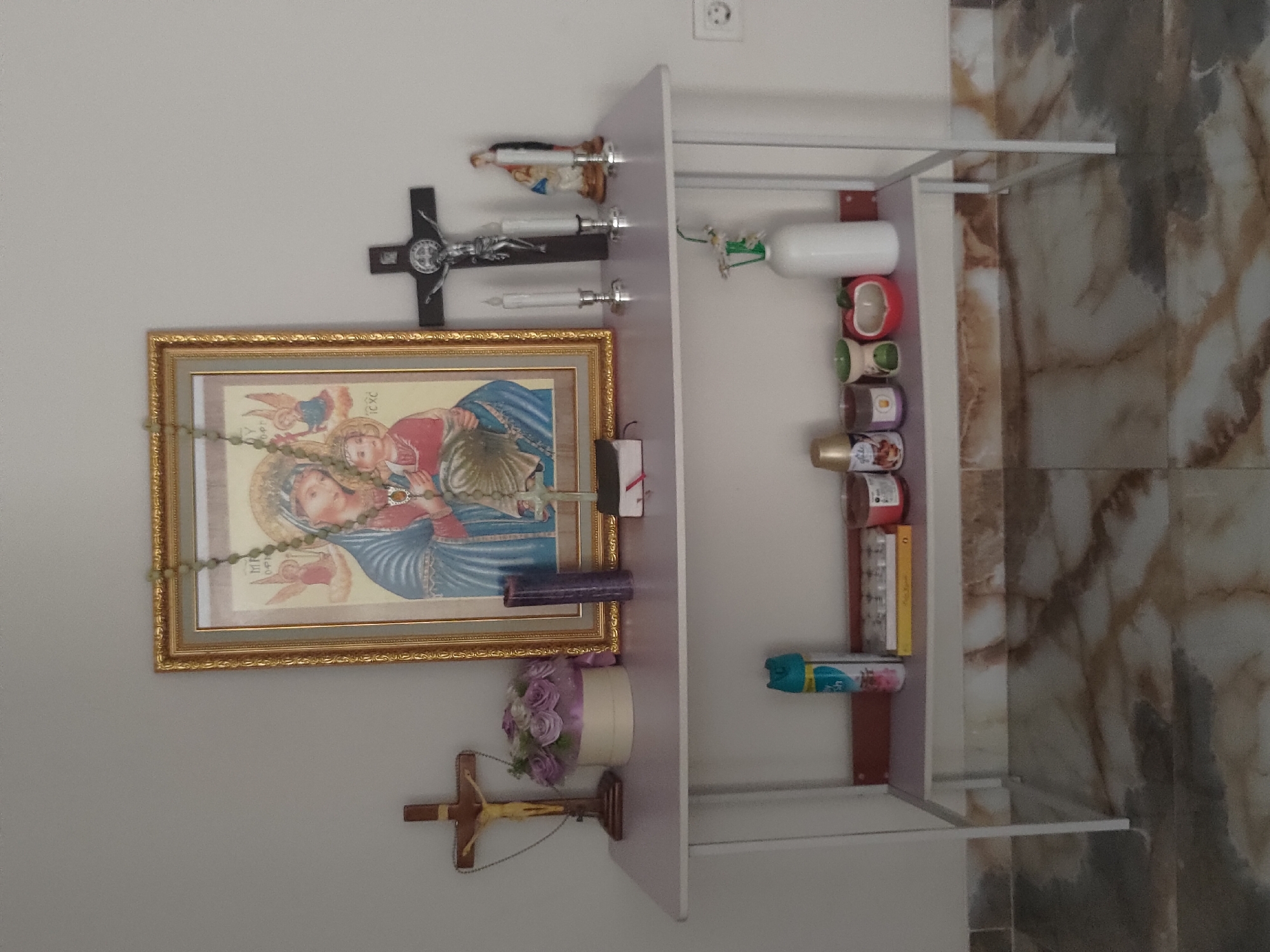 This is a close-up, eye-level shot of a shelf displaying religious items. The shelf is made of a light purple material with white metal legs and is placed against a light gray wall. On the top shelf, a framed religious icon of Mary and Jesus is prominently displayed, adorned with a green rosary. To the left of the icon is a wooden crucifix, and to its right are two white candles in silver holders and a dark crucifix with a silver figure of Jesus. A statue of a saint is also present. A dark purple candle stands next to the icon.
The lower shelf holds several items including a can of air freshener, a yellow box with small items, several candles of various sizes and colors, and a white bottle with small white flowers. The floor is covered in marble tiles with brown and white patterns. A white electrical outlet is visible on the right side of the wall. The scene appears to be indoors. The location is described as Depok, Indonesia.
