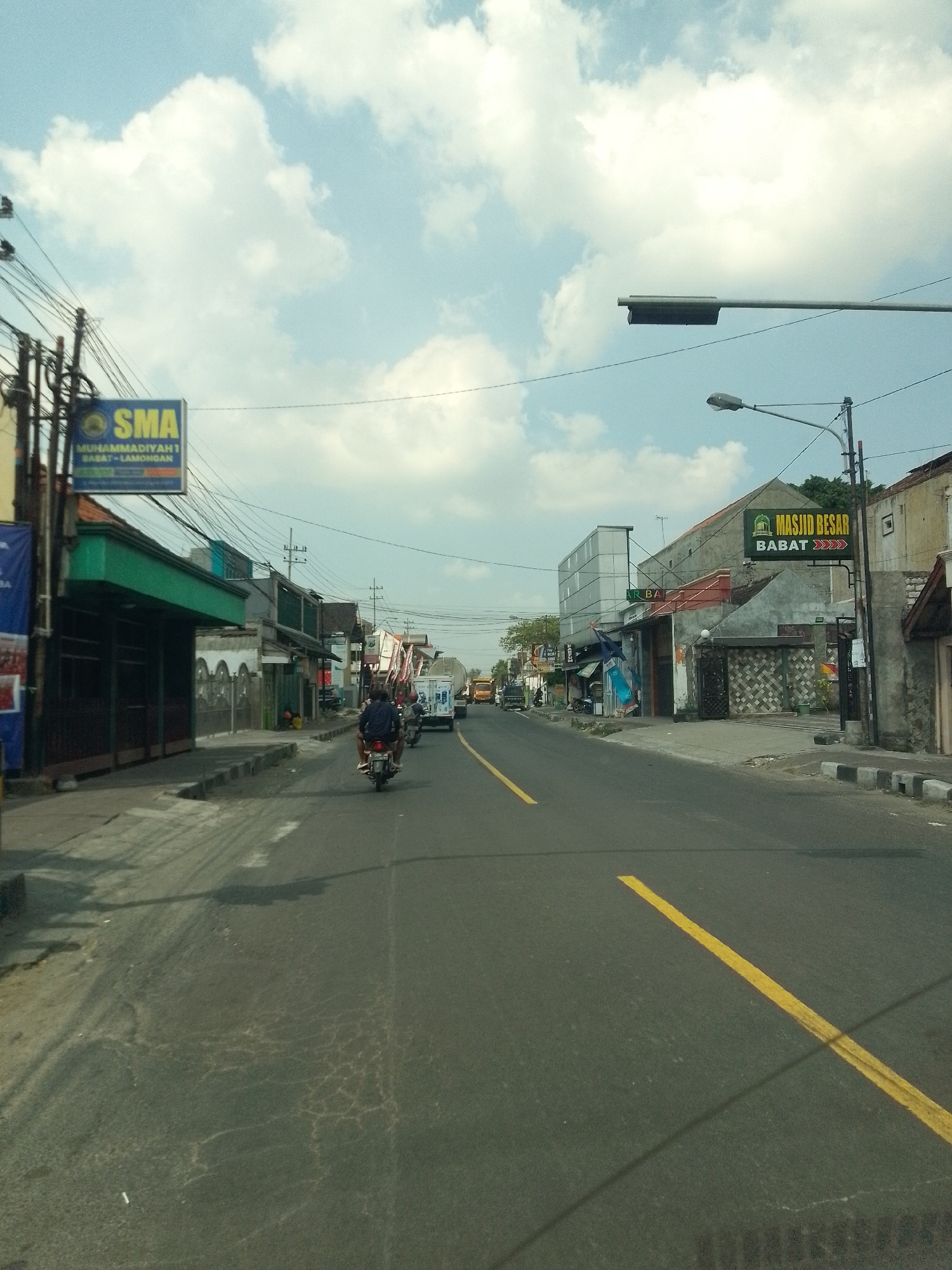 The image depicts a street scene in Babat, Indonesia. A road with a yellow dividing line stretches into the distance. On the left side of the road, a sign indicates "SMA MUHAMMADIYAH 1 BABAT - LAMONGAN," suggesting a high school. Further down the street, a sign for "MASJID BESAR BABAT" identifies a large mosque. Several buildings, including shops and what appears to be a mosque, line the street. Traffic is present, with a motorcycle carrying two individuals in the foreground, and a truck and other vehicles further ahead. Utility poles with numerous wires are visible throughout the scene, as are streetlights. The sky is partly cloudy. The overall impression is of a moderately busy urban environment.
