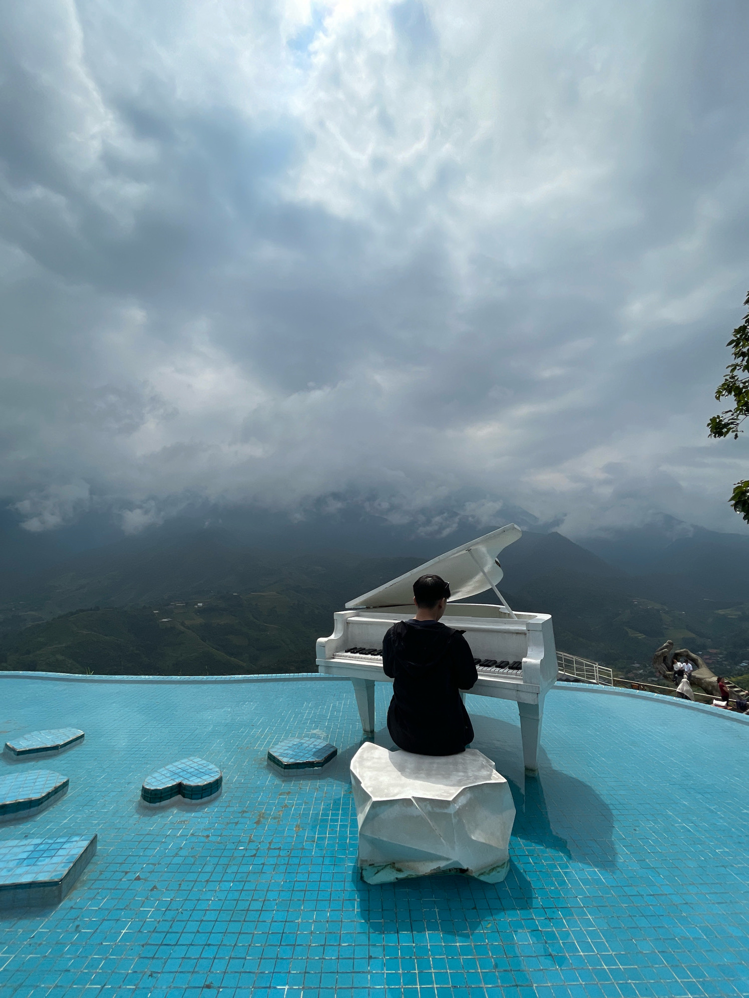 The image shows a person playing a white grand piano on a tiled, blue swimming pool overlooking a mountainous landscape in Sa Pa Ward, Vietnam. The sky is filled with heavy, grey clouds, with patches of light breaking through in the upper left corner. The piano is positioned on a stone block, with the player seated behind it, facing away from the camera. The swimming pool has several heart-shaped tiles or small islands floating in it. In the background, the mountains are covered in lush green vegetation, with some visible structures and terraced fields. A large hand-shaped structure with people on it is visible in the far right background, near a railing.