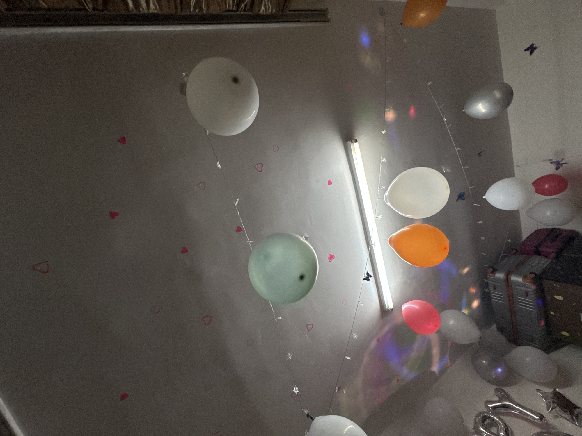 The image shows a room decorated with balloons of various colors (white, orange, silver, pink, green) and shapes. Some balloons are tied with strings and adorned with small lights or decorations. The walls are light-colored and feature small heart-shaped stickers. A fluorescent light fixture is visible on the wall. In the background, there are pieces of luggage or storage containers. The location is not evident from the image, and there is no indication of Zhongtun, China.