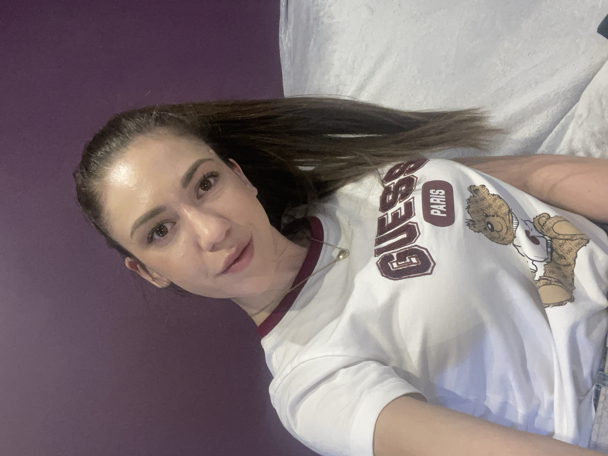 The scene depicts a female individual in an indoor environment, located in Sofia, Bulgaria. The subject, with dark brown hair pulled back into a ponytail, is positioned facing the camera, her head tilted slightly to her left. She wears a white short-sleeved t-shirt with a maroon collar band. The t-shirt features a graphic print consisting of the maroon text "GUESS PARIS" above a cartoon teddy bear figure detailed with a "G" pattern. A gold-colored necklace with a small, pear-shaped pendant is visible around her neck. The background is divided, with a solid purple wall or surface occupying the upper left quadrant and a textured white or light grey fabric, consistent with a blanket or upholstery, filling the right side of the frame.