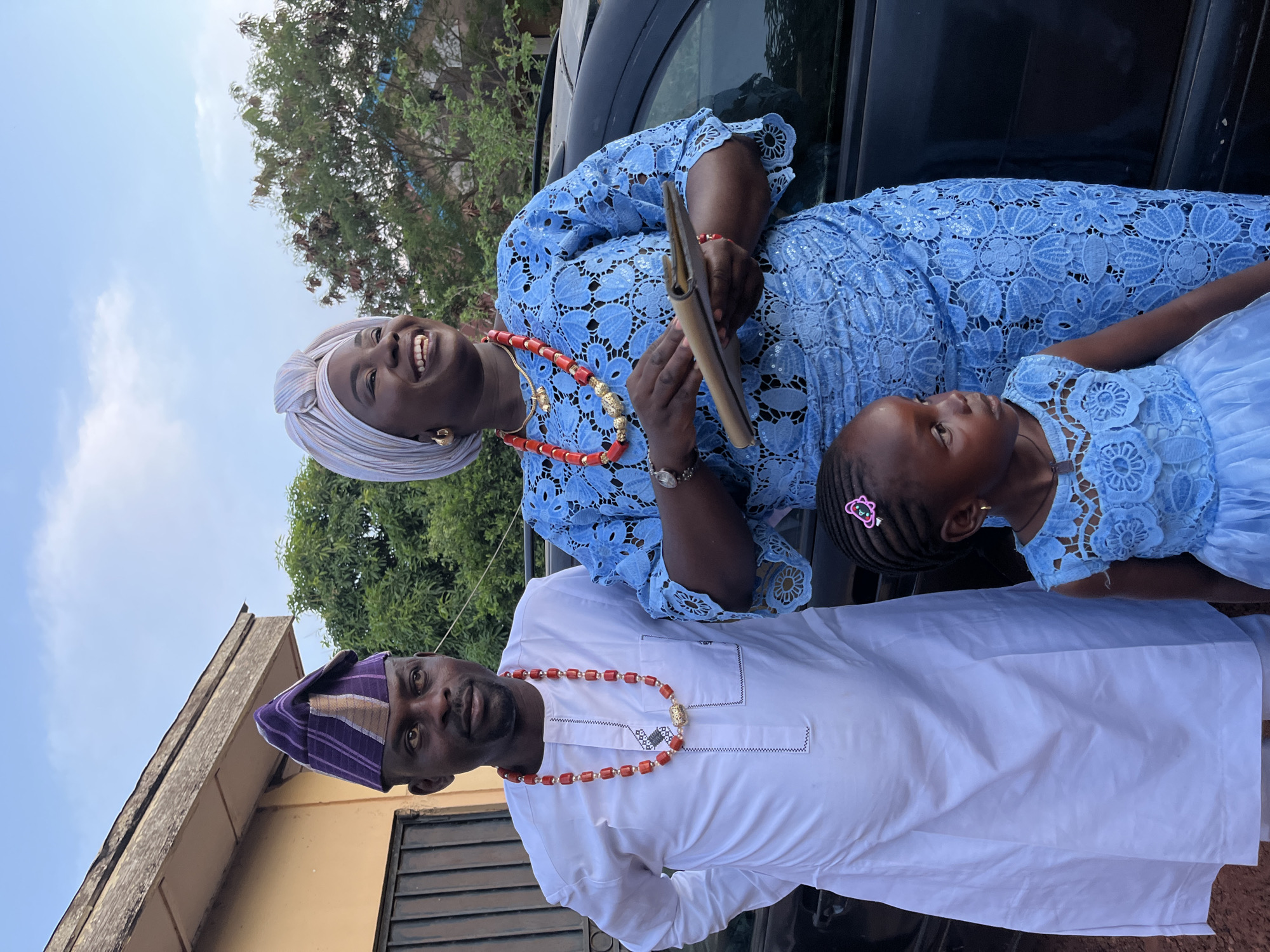 Three people pose together in an outdoor setting in Rafin Bauna, Nigeria. An adult woman wearing a light blue lace dress, a light-colored head wrap, and a red beaded necklace smiles broadly. She holds a brown wallet or clutch in her hands. A young girl wearing a matching light blue lace dress with a pink hair clip stands beside her. An adult man, wearing a white traditional shirt and a purple-striped cap, stands near them, also wearing a red beaded necklace. They stand in front of a dark-colored car, with trees visible in the background. A building is partially seen in the lower left corner. The sky above is light blue with some clouds.