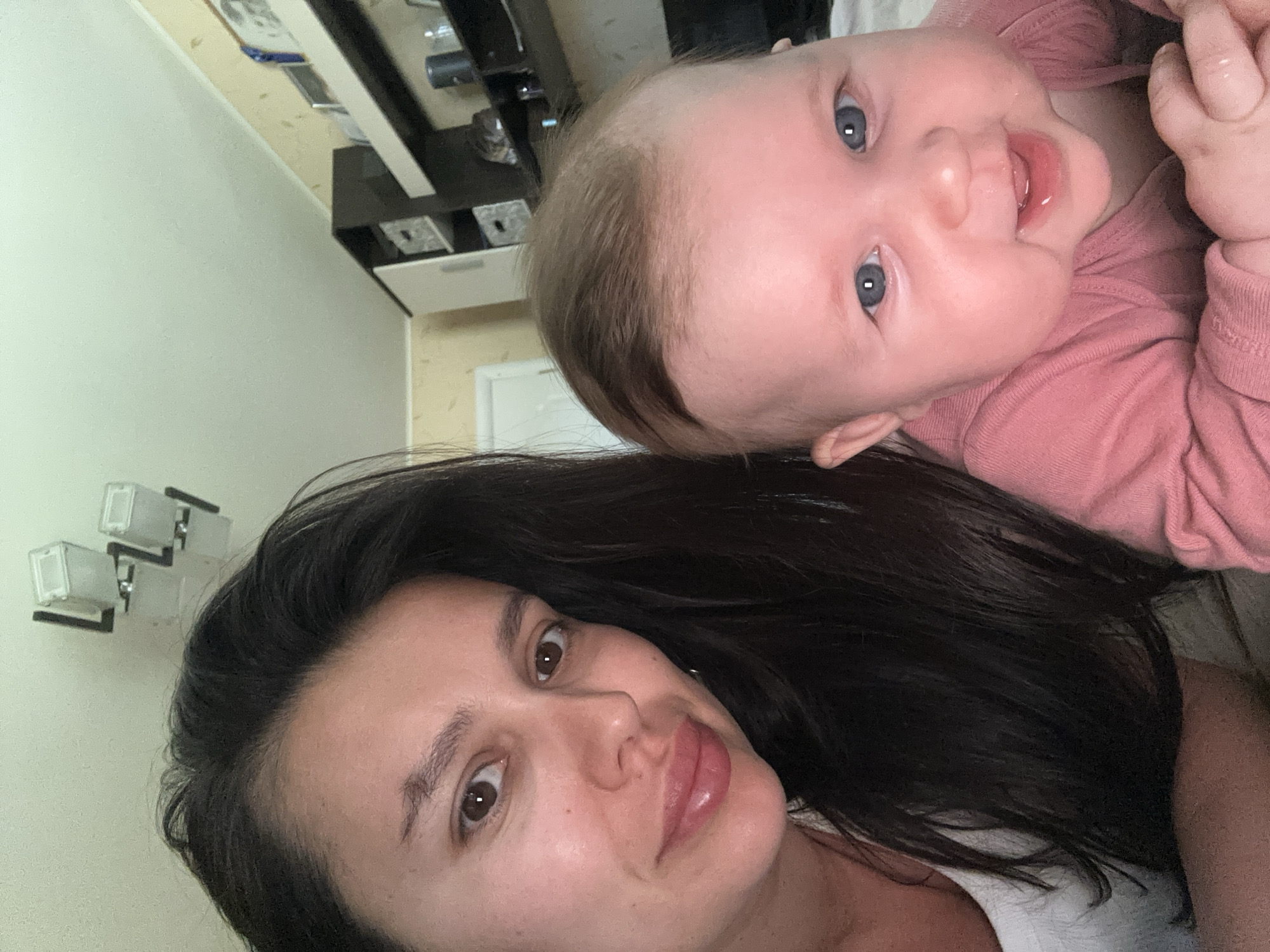 A selfie captures a woman with dark hair and a baby in a pink outfit. The woman is fair-skinned with brown eyes and full lips. The baby has fair skin, light brown hair, and blue eyes. They are presumably indoors, with a wall-mounted light fixture and a cabinet visible in the background. The exact location is the city of Chaikovsky, Russia.