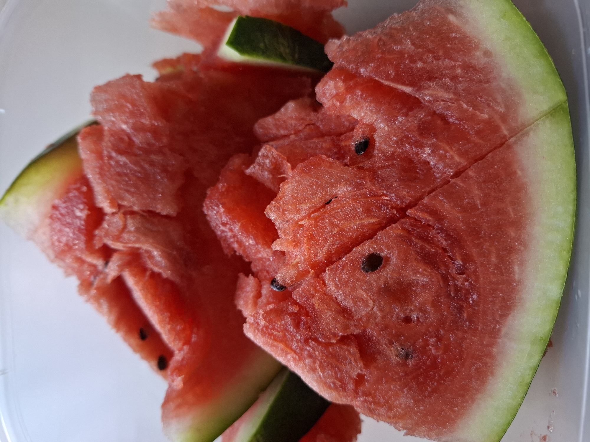 A close-up, overhead shot displays several triangular slices of watermelon artfully arranged in a clear plastic container. The bright red flesh of the watermelon is textured and juicy, with visible black seeds scattered throughout. The rind of the watermelon is a vibrant green, transitioning to a pale white inner layer. The background is a soft, blurry white, indicating a plain surface or wall. The lighting is bright, casting subtle shadows on the fruit, highlighting its freshness. The scene appears to be a still life of a refreshing summer treat. The image does not contain any elements that would indicate a specific location in the city of Tyumen, Russia, or any details about people or actions.