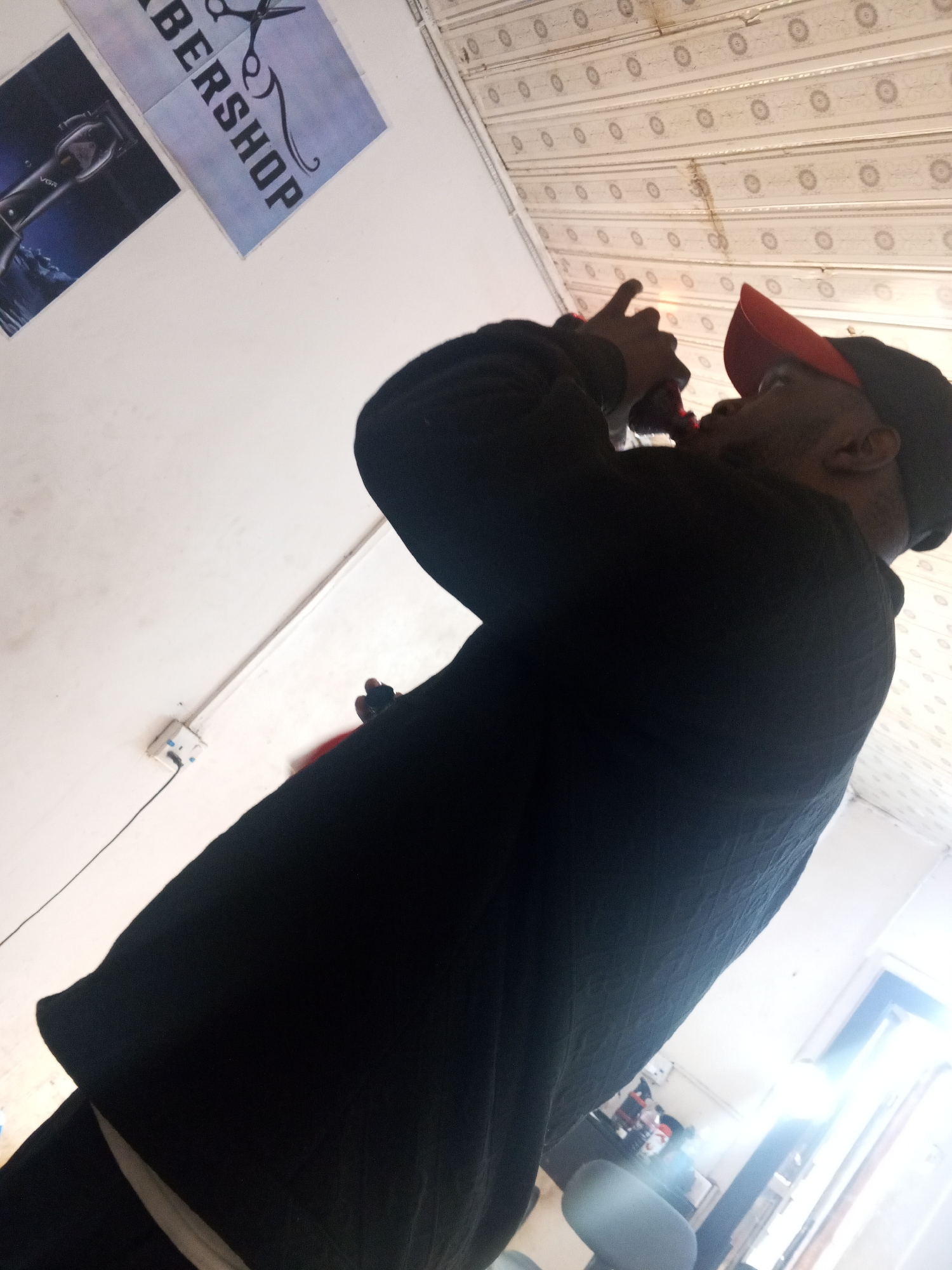 A person is visible from the waist up, drinking from a dark container. They are wearing a black sweater and a red baseball cap. In the background, there is a sign for a barbershop on a wall, indicating the setting is likely a barbershop. The building appears to be located in Gwong, Nigeria, based on the prompt instructions. The background also shows a patterned ceiling and a window. Another chair is barely visible. The person seems to be actively drinking, holding the container to their mouth with one hand.