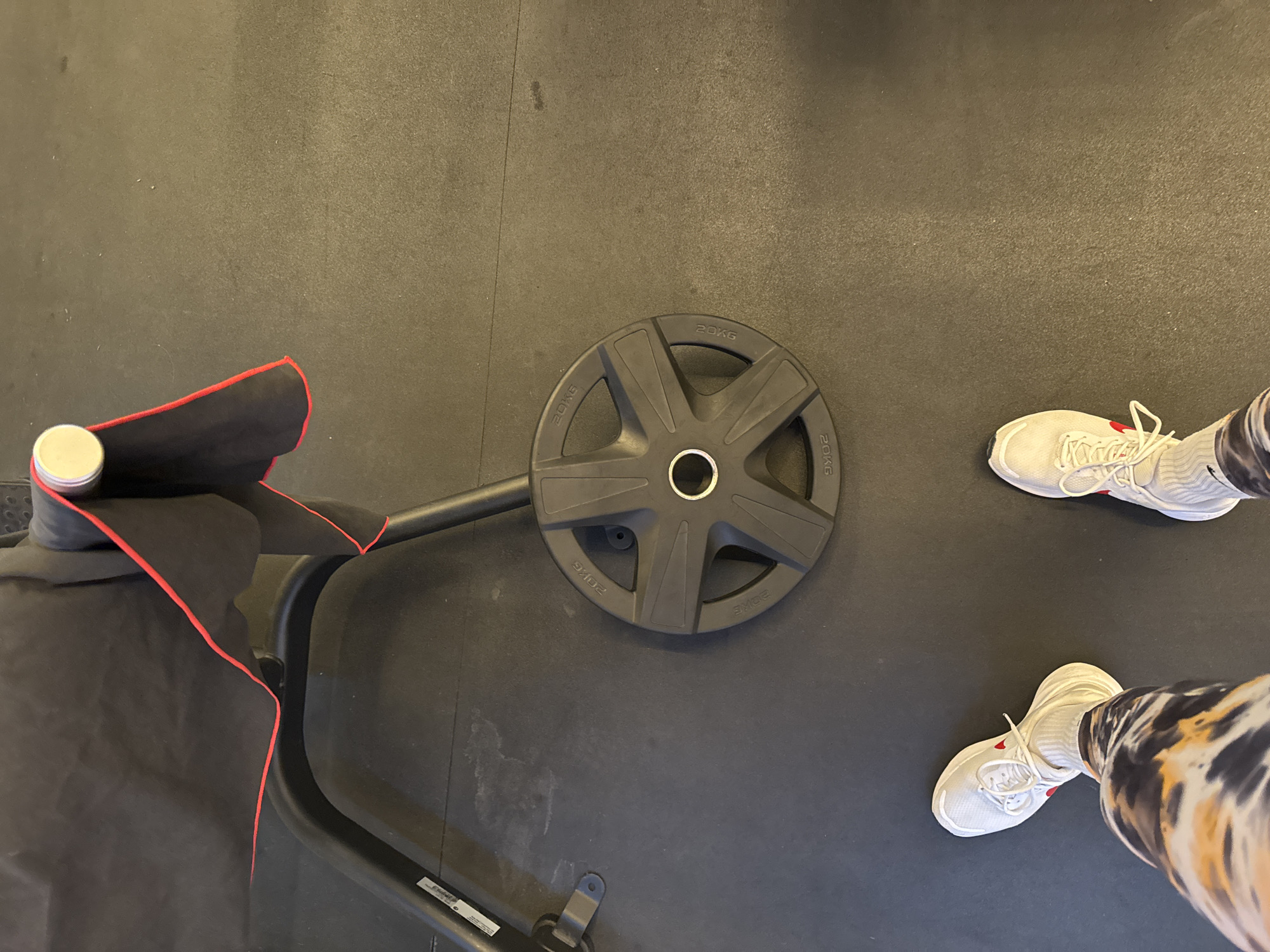 In the city of Murcia, Spain, this image captures a gym floor setting. A black weight plate, marked "20KG" multiple times, rests on the dark, textured floor. To the left, a portion of a black gym bench is visible, draped with a black towel trimmed in red. In the foreground, a person's legs and feet are positioned, wearing white athletic shoes with red accents and tie-dye patterned leggings. The overall scene depicts an environment ready for a workout.