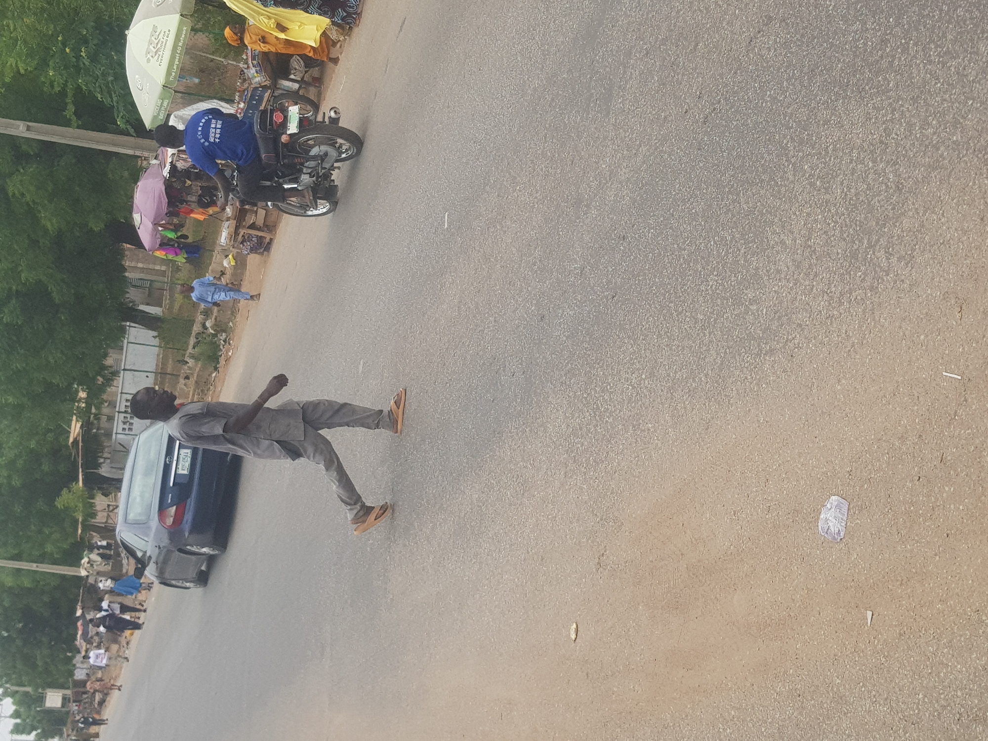 The image is of a street in Dabawa, Nigeria. A man is walking in the street, and a blue car is parked on the side of the road. A motorcycle is parked near a vendor with a blue shirt that has Chinese words on it. Trees and foliage line the left side of the frame, and a group of people can be seen gathered on the side of the road behind the car. The road itself is made of asphalt and has some trash and debris scattered on it.