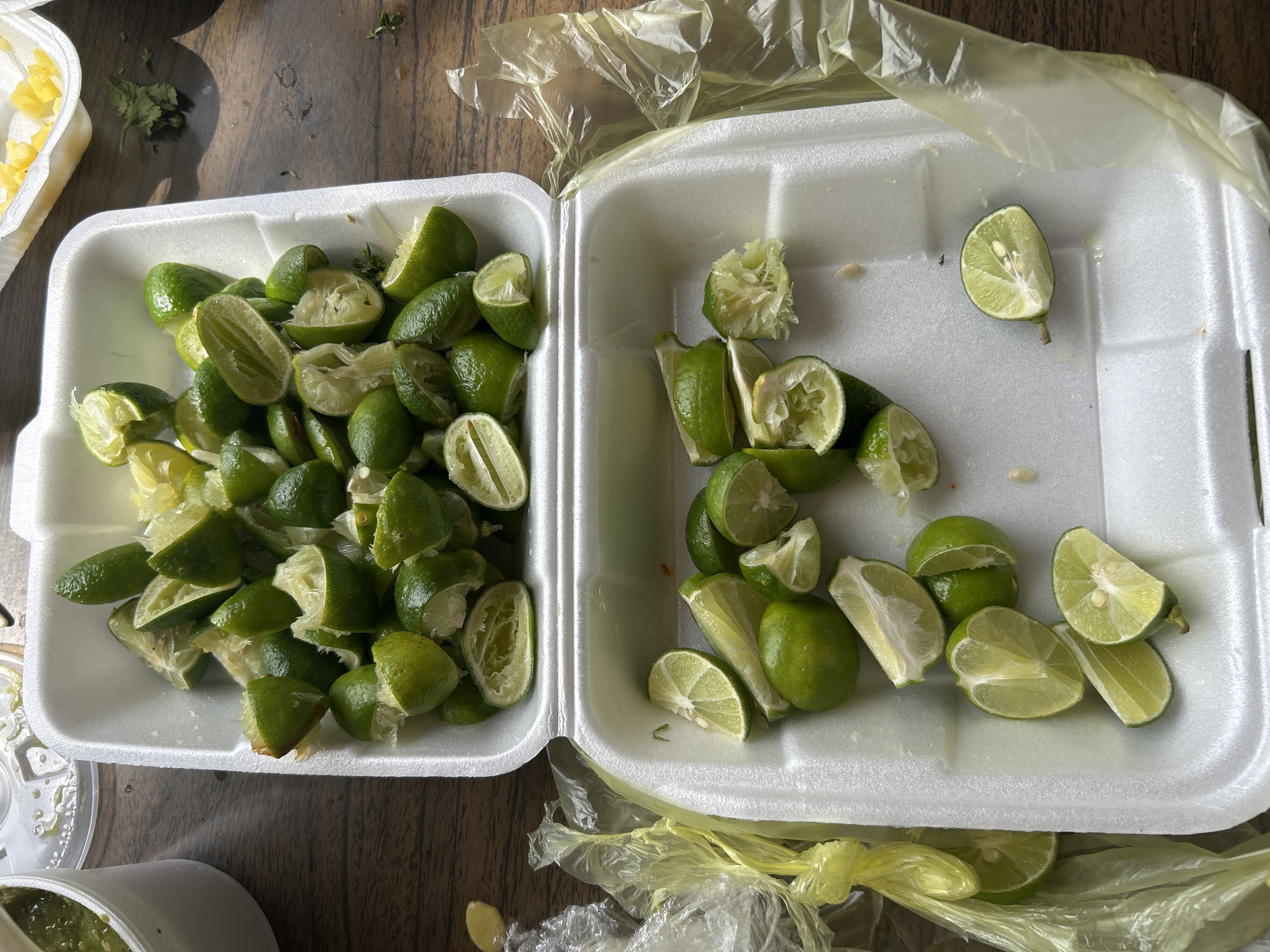 The image shows two opened styrofoam containers filled with squeezed lime halves on a dark wooden surface. The left container is densely packed with lime halves, while the right container has fewer, more spread-out lime pieces. A plastic bag is present around the right container. A small container with yellow contents sits to the left of the container with limes. The location is Unknown, Unknown.