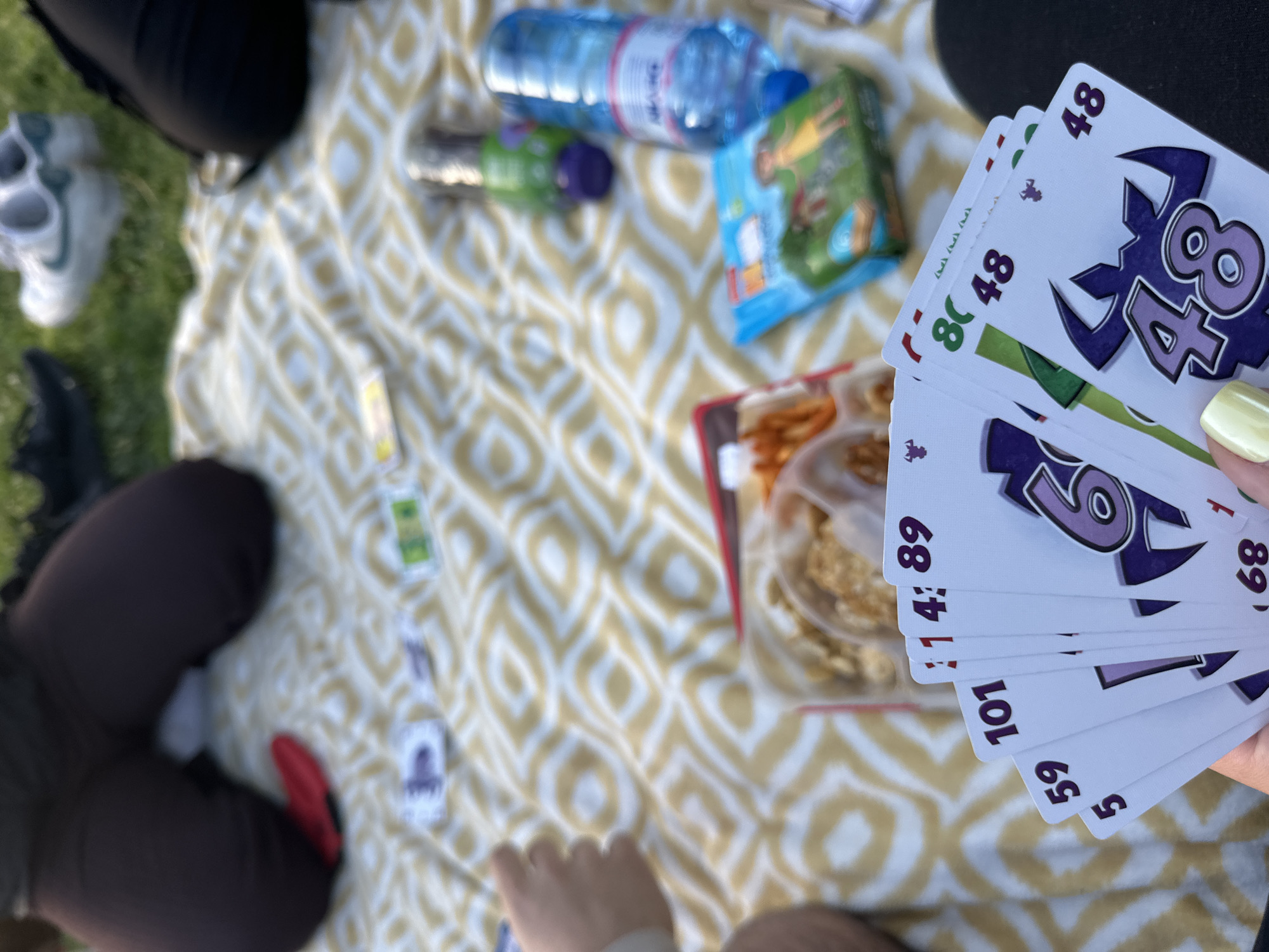 A picnic scene unfolds outdoors, likely in a park. A patterned blanket is spread on the grass. Several people are present, although only parts of their legs and clothing are visible. A hand holds playing cards with large numerals on them; the visible cards display the numbers 59, 101, 114, 89, 69, and two 48s. A partially visible snack container, a sealed pack of treats, a water bottle, and another drink bottle sit on the blanket. Additional scattered cards are nearby. The location is assumed to be Sofia, Bulgaria.