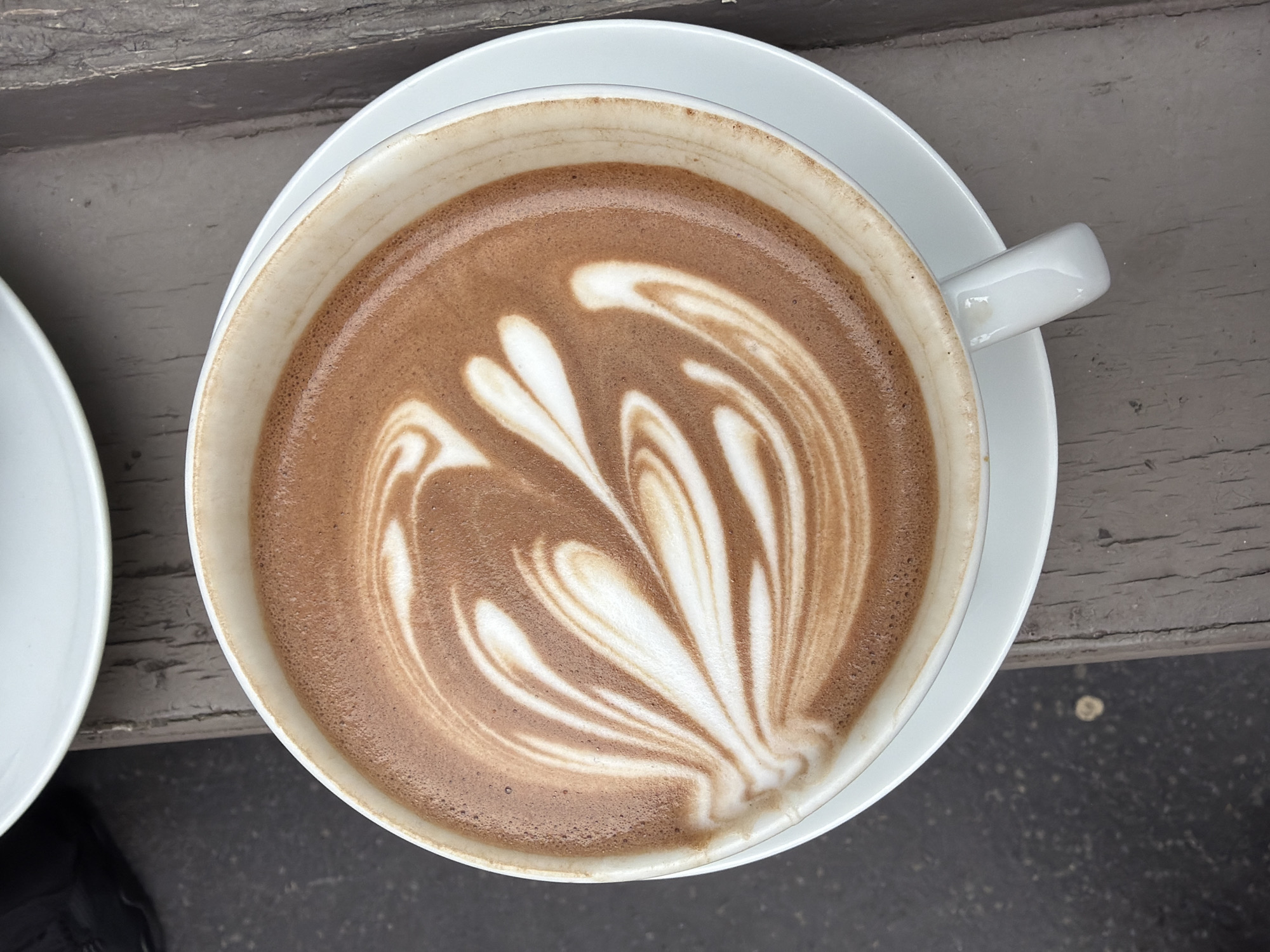 A top-down shot presents a cup of latte art on a white saucer, resting on a grey wooden surface. The latte art features a leaf-like design in white, contrasting with the brown coffee base. The cup is white with a matching white handle. Part of another white saucer and a dark shoe are visible in the lower left corner. The image appears to have been taken outdoors on a weathered wooden table, potentially in a cafe setting. There is no explicit indication of Paris, France in the image.