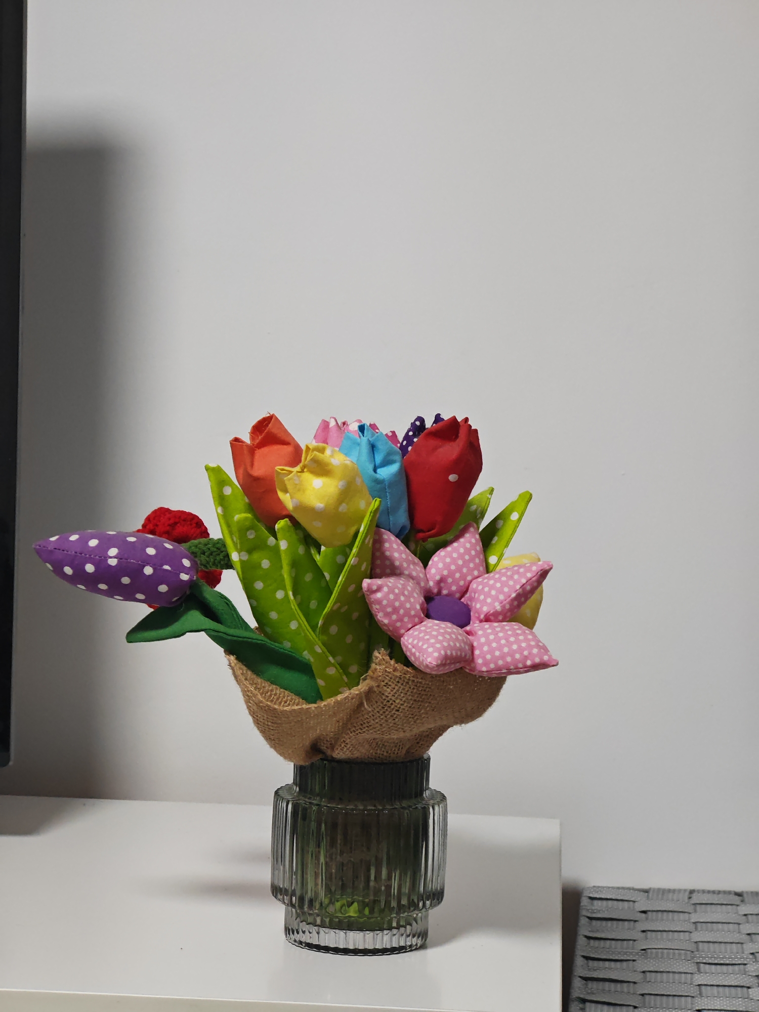 A multicolored bouquet of artificial fabric flowers is displayed in a dark, ribbed glass vase, resting on a white flat surface against a plain white wall. The bouquet consists of various fabric flowers, including red, yellow, blue, and light pink tulips, many featuring white polka dots. Green fabric leaves, some also with white polka dots, are interspersed. A prominent five-petal flower, light pink with white polka dots, has a central purple button. A purple, pod-shaped fabric flower with white polka dots and an adjacent red crocheted element is positioned on the left side of the bouquet. The base of the flower arrangement is wrapped in a light brown, coarse, burlap-like material. To the far left, a dark vertical edge of an unknown object is visible. To the far right on the white surface, a section of a gray, horizontally woven textured object is present. No individuals are depicted. The visual evidence within the image does not provide sufficient information to specify a precise location within the city of Shumen, Bulgaria.