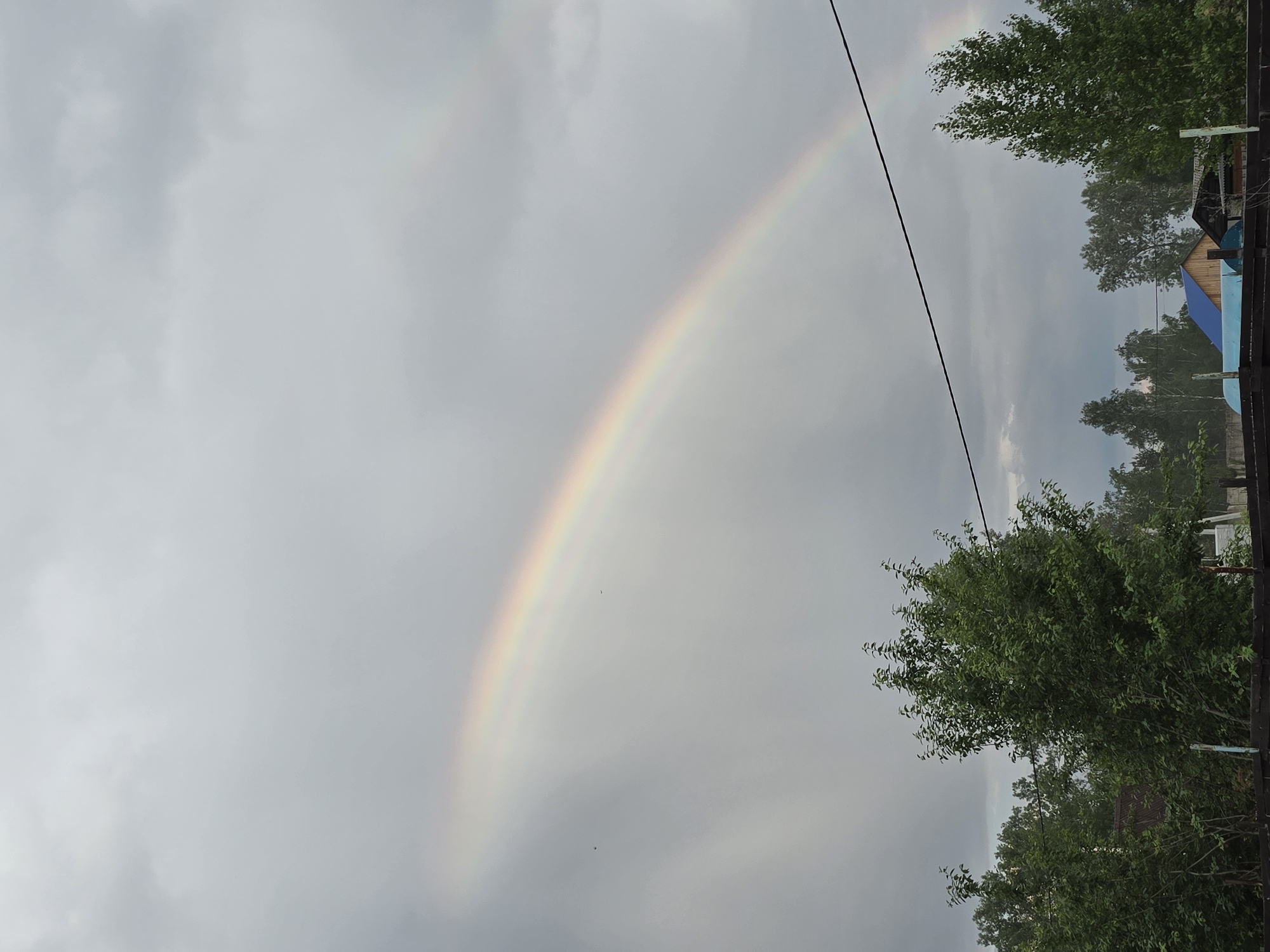 A wide-angle shot captures a serene rural landscape under a cloudy sky. A vibrant rainbow arcs across the grey expanse, its colors softly diffused by the overcast conditions. In the foreground, lush green trees form a dense border, their leaves rustling in a gentle breeze. Below the trees, glimpses of rustic houses and fences suggest a quiet village life. The scene is devoid of human presence, creating a sense of peaceful solitude.