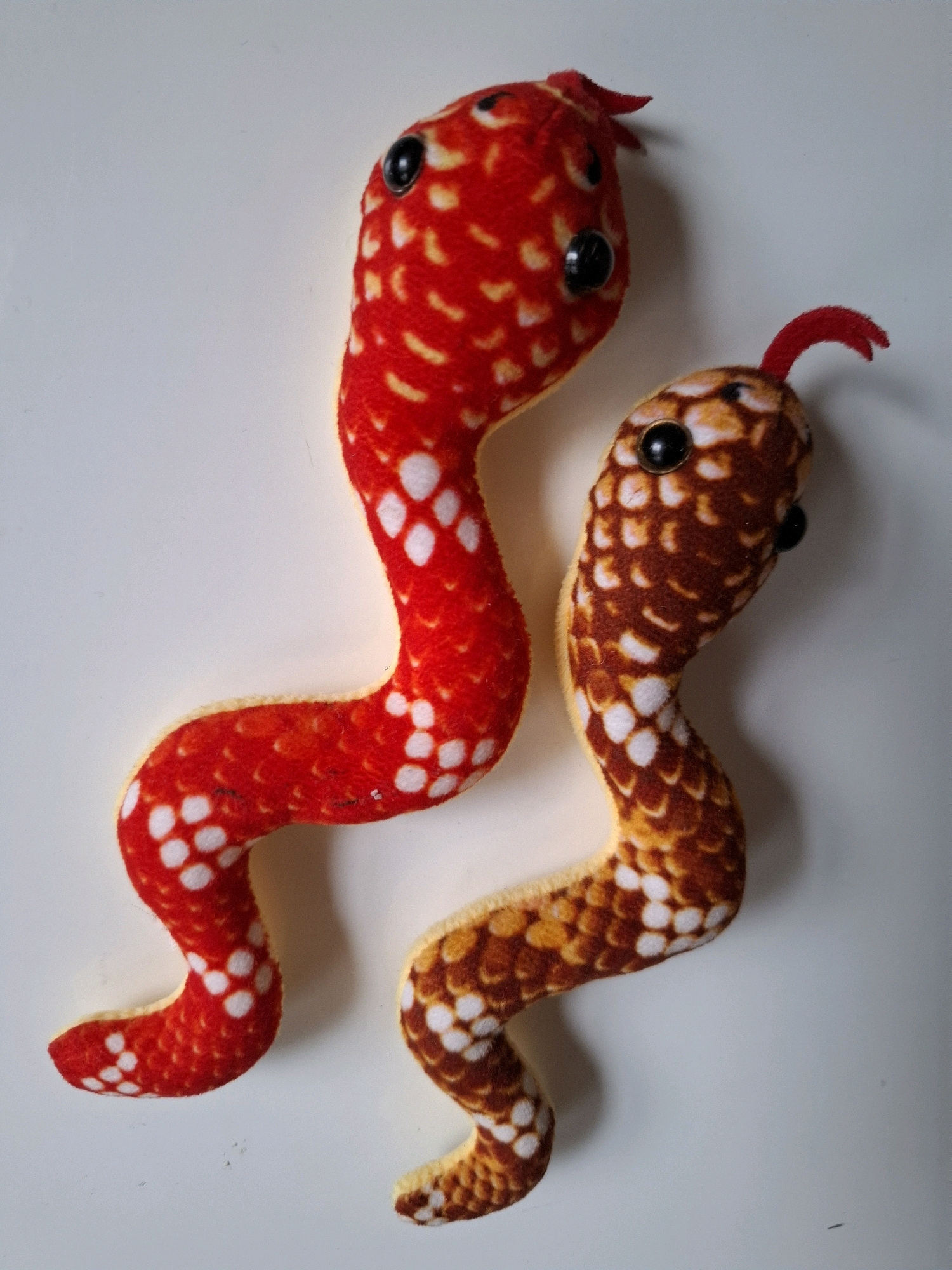 Two plush toy snakes are lying on a white surface. The snake on the left is red with white and yellow spots. The snake on the right is yellow with brown and white spots. Both snakes have black plastic eyes and red felt tongues. The lighting suggests a soft, diffused light source from the top right. The scene does not depict any identifiable landmarks or elements that would indicate a specific location in the city of Tyumen, Russia.