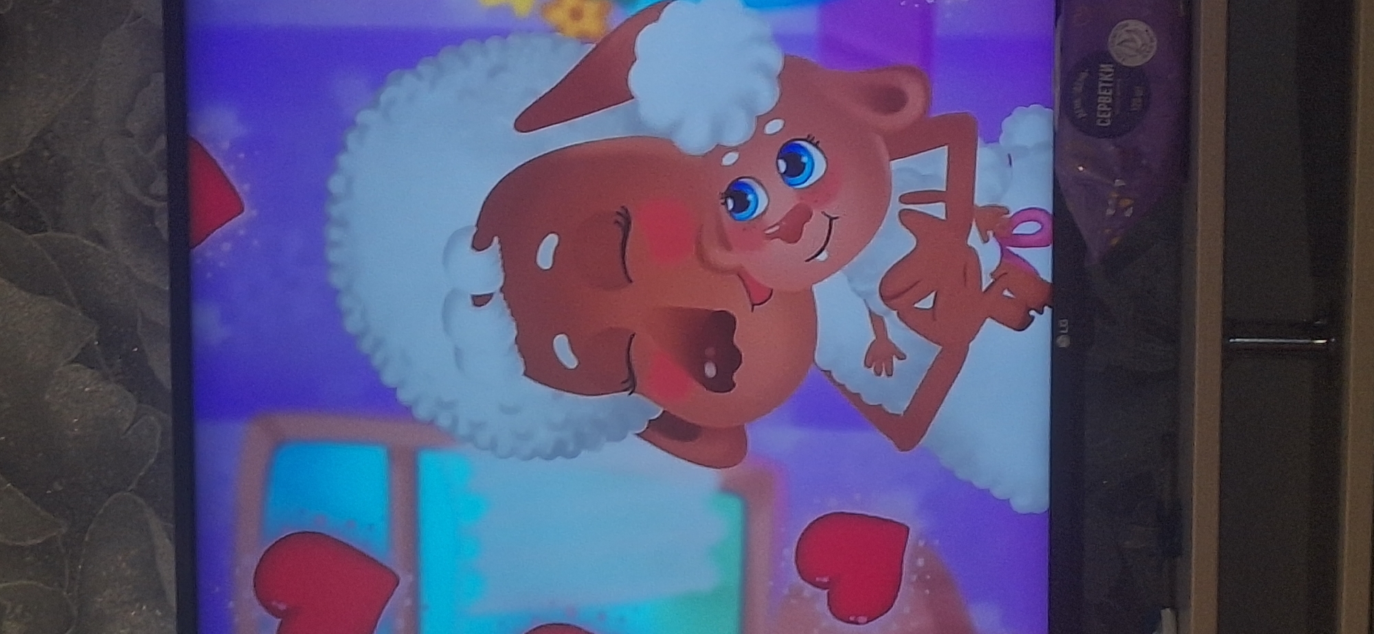 The image shows a television screen displaying an animated scene of two sheep characters. The larger sheep, with white curly wool and brown skin, is embracing a smaller sheep. The smaller sheep, also brown-skinned with blue eyes and wearing a red Santa hat, is nestled in the larger sheep's arms. The background is a soft purple with scattered red hearts and some yellow flowers on the right. A shelf with a package of wet wipes labeled "СЕРБЕТКИ" (servetki, meaning "wipes" in Russian) is visible below the television screen. The location is not specified, and there is no information about Bila Tserkva, Ukraine.