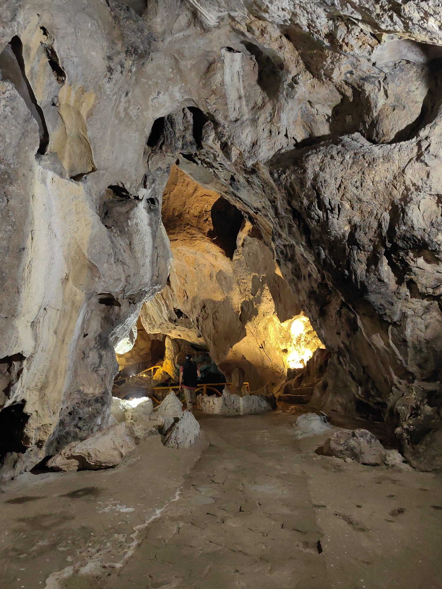 This image depicts the entrance to a cave. The cave walls are made of light-colored rock with darker patches and are rough and uneven. The entrance is partially lit by an orange glow from within the cave. A person wearing a dark vest and light pants stands inside the cave, near a wooden railing. The floor of the cave is a mix of dirt and rock. This cave is in Lạng Sơn, Vietnam.