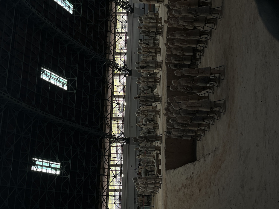 The image displays an archaeological excavation site, specifically a large hall housing the Terracotta Army, located in the Lintong District, China. Hundreds of life-sized terracotta warrior and horse statues are observed in military formations within extensive, unearthered trenches. The foreground features multiple rows of standing terracotta figures, many stabilized by visible metal support frames. Beyond these, additional terracotta soldiers and horses populate further excavated pits, some appearing in various stages of restoration or still partially encased in the reddish-brown earth. The overhead structure of the building is characterized by a dark, visible framework of steel trusses, punctuated by several rectangular skylights or overhead lighting fixtures. A long, horizontal band of grid-patterned windows is visible along the distant wall, allowing for natural light penetration. Faint human figures, consistent with visitors or personnel, are discernible observing the formations in the background. The scene is dimly lit by a combination of natural and artificial light sources, casting shadows across the excavated areas.