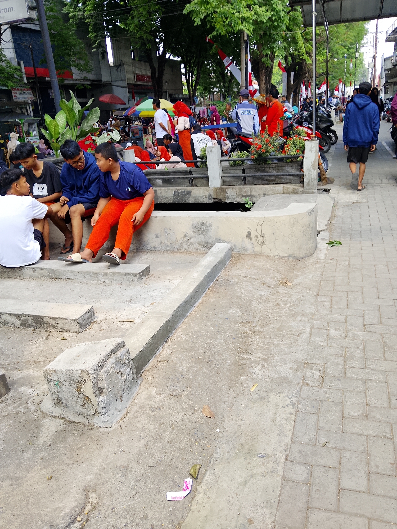 The image depicts a street scene in Kertosono, Indonesia, likely near a public area or market, given the presence of vendors and a busy atmosphere. The foreground shows a group of young males sitting on concrete structures, some of them appearing to be conversing. One individual on the right is wearing bright orange trousers, while others are in casual t-shirts and shorts. Behind them, a variety of people are visible, including individuals in uniform and others in casual attire. Motorbikes are parked along the side of the street, indicating a mode of transportation common in the area. The background features buildings, trees, and decorative flags, suggesting a festive or community event. The overall environment is urban, with paved walkways and a mix of natural and man-made elements.
