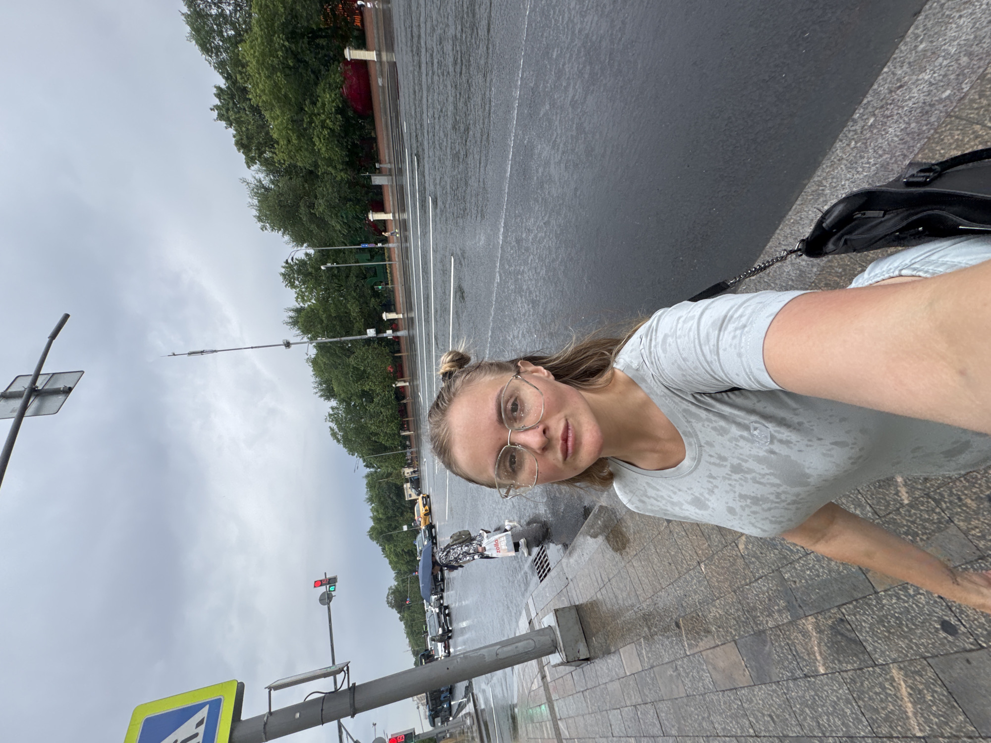 A woman is taking a selfie on a rainy day in Moscow, Russia. She is wearing glasses and a gray t-shirt. The road and sidewalk are wet, and it is raining heavily. In the background, there are cars, buses, and trees. A pedestrian crossing signal is visible, showing a red light. The woman is holding a black purse.