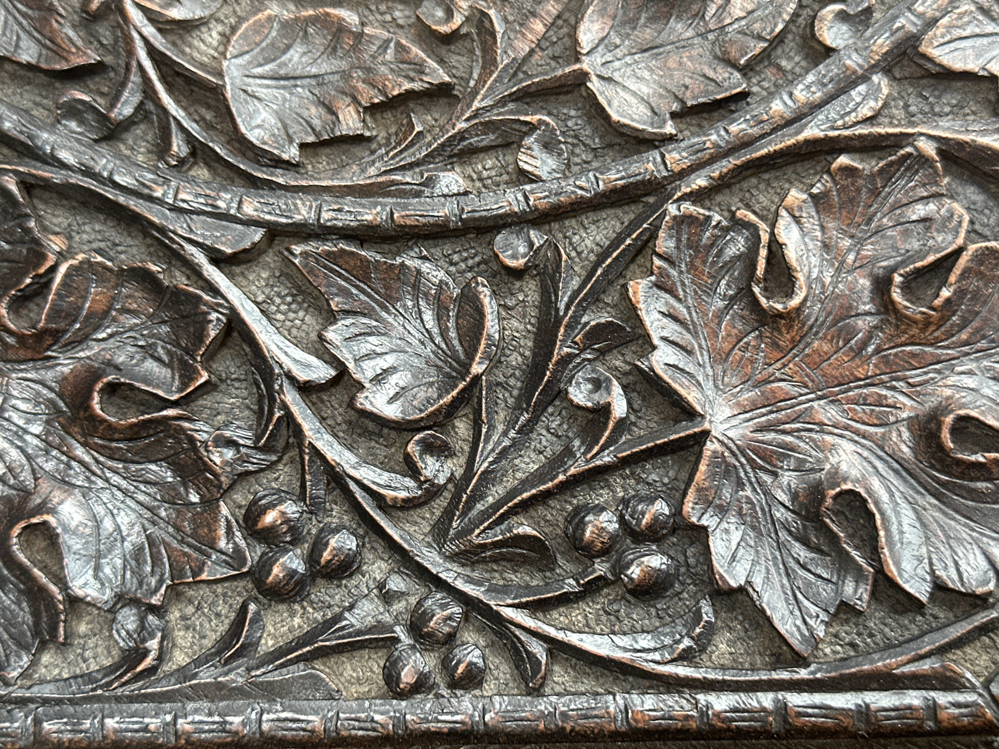 This is a close-up photograph of a section of intricately carved wooden ornamentation. The pattern consists primarily of leaves, vines, and small round objects resembling grapes. The wood is a dark brown color, with variations in tone suggesting shadows and highlights that accentuate the depth of the carving. The carving style is detailed and appears to be from a traditional or historical context.
Given the question's request to include Tunbridge Wells, United Kingdom, the carving could potentially be a detail from architectural features within the town, or decorative elements found on antique furniture or artwork residing there. However, there is no context from this image that would identify it as originating from that specific location.